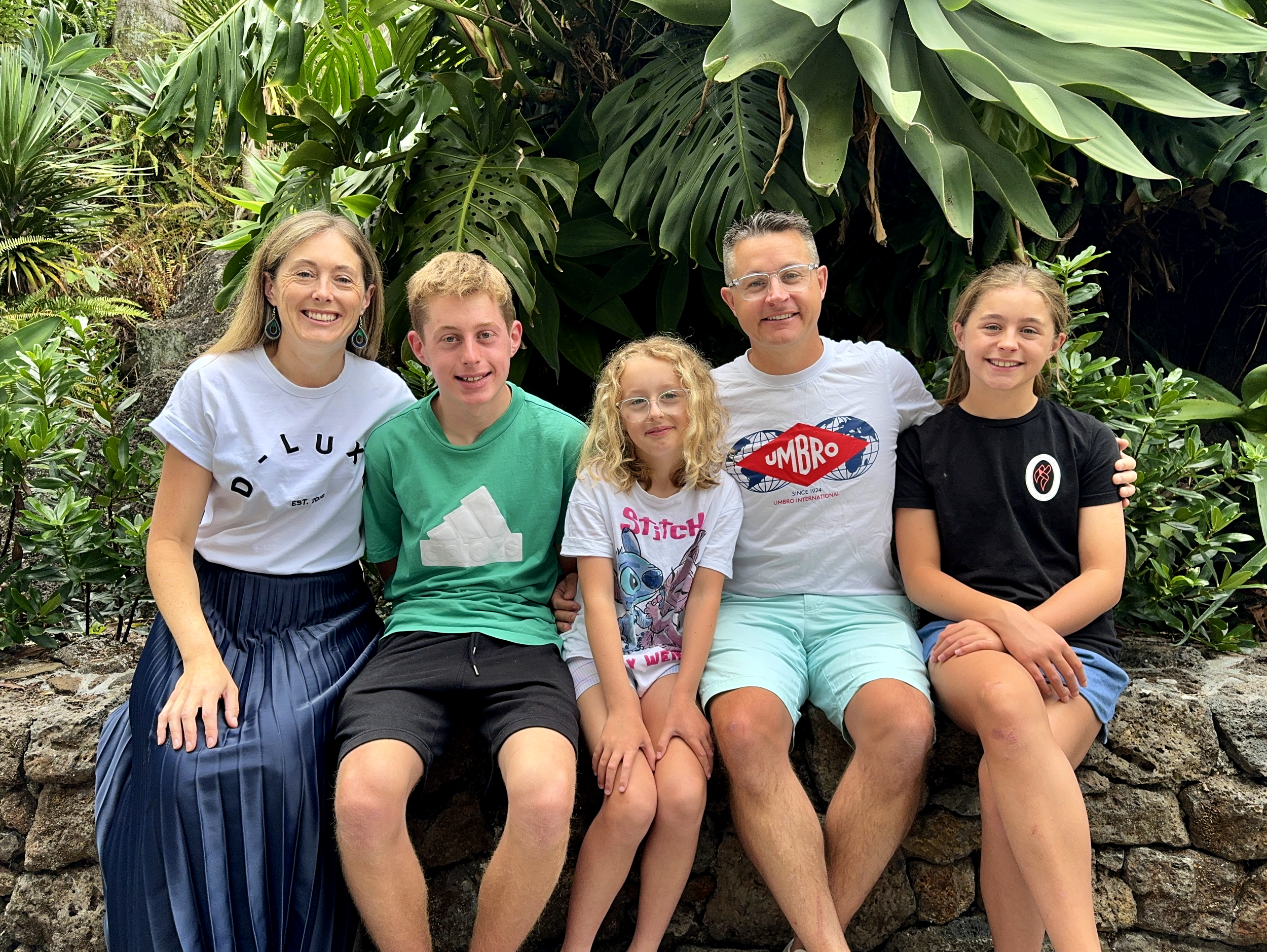 A group of five people sitting outdoors in front of large green tropical plants, smiling at the camera.