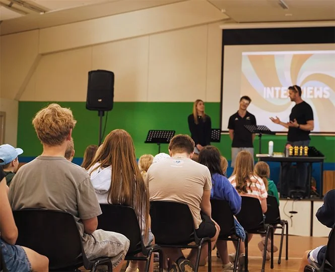 A group of children seated in a classroom or auditorium, listening to three presenters standing at the front near a large screen displaying the word 'INTERVIEWS' with colorful graphics.