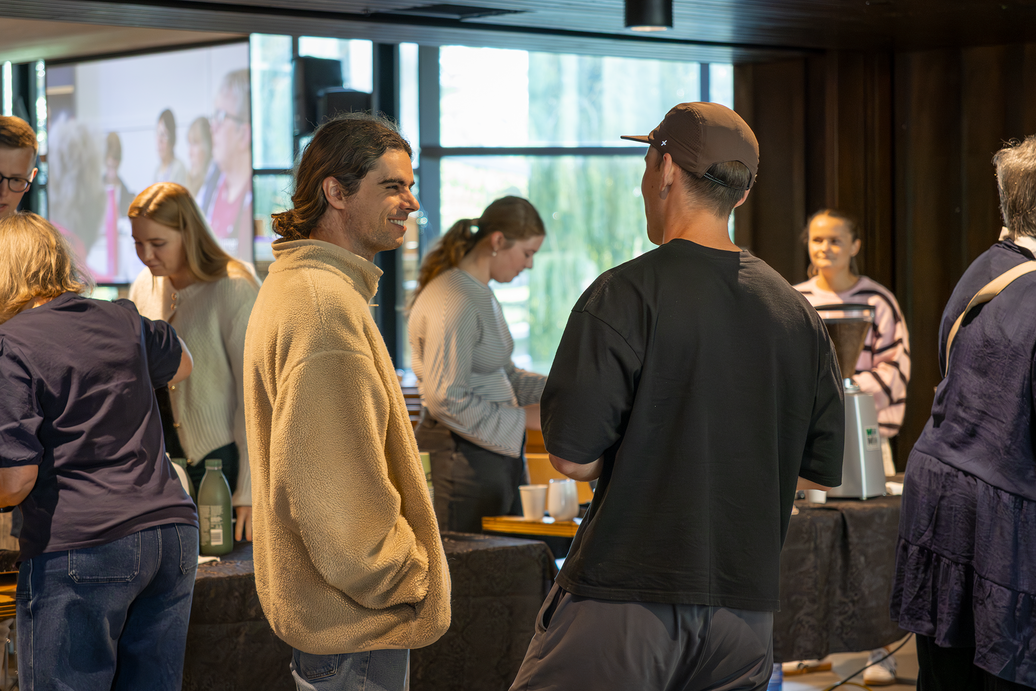 People at an indoor event, engaging in conversation near a counter with beverages and a coffee machine.