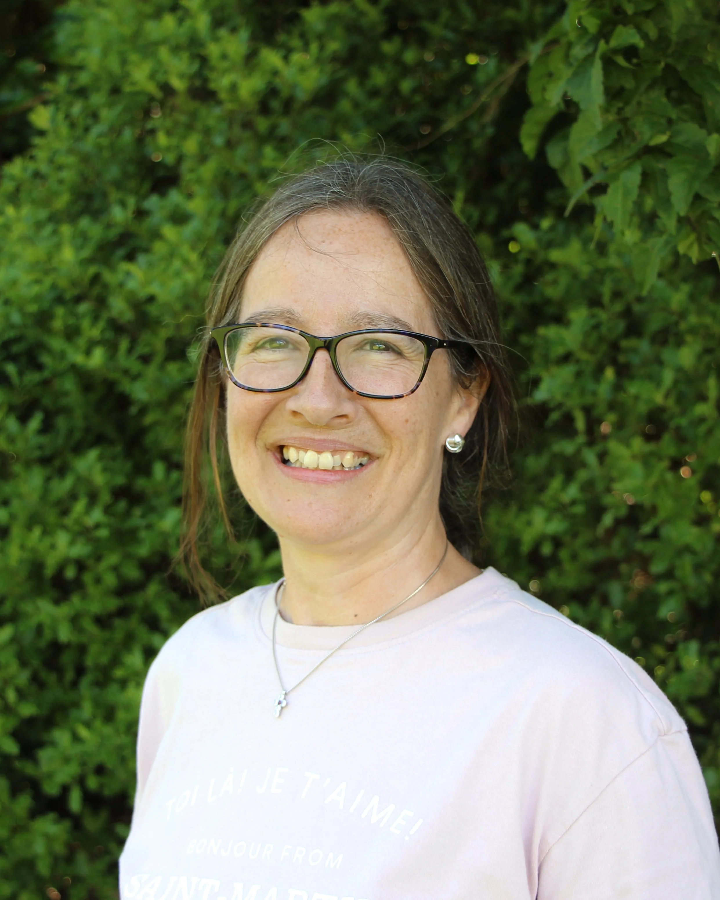 Close-up of a smiling woman with glasses, wearing a white T-shirt and a necklace, standing in front of green leafy bushes.