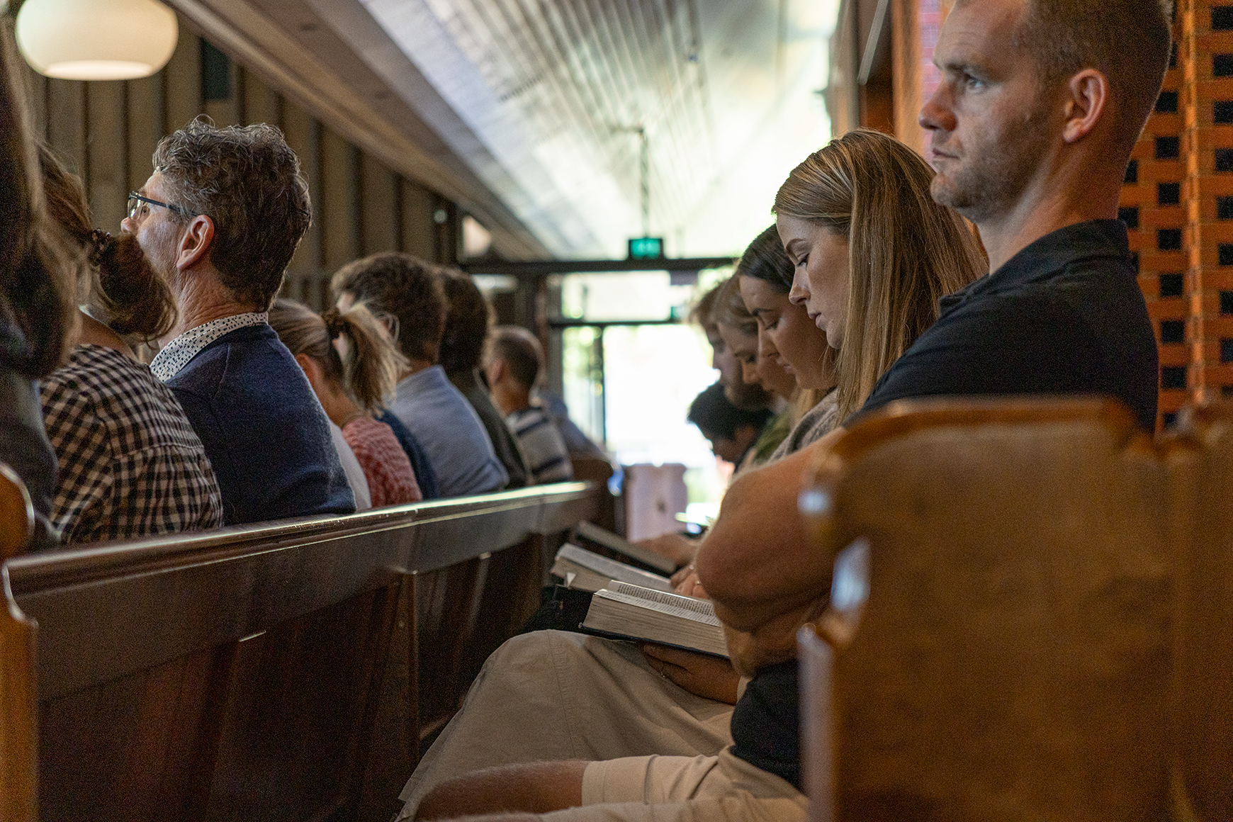 People sitting in pews during a church service or event, some reading books or looking straight ahead, inside a church with wood-paneled walls and high ceiling.