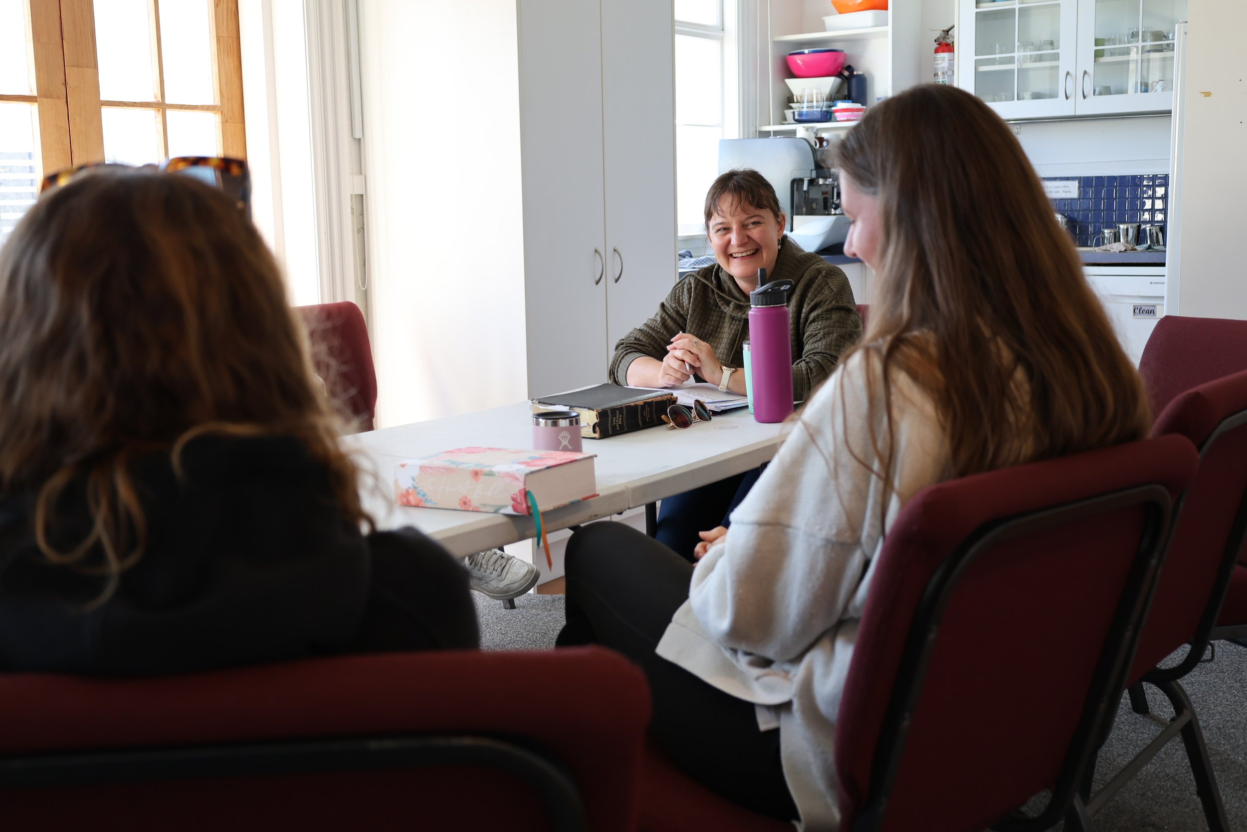 A woman with short hair smiling and sitting at a table during a meeting with three other women inside a kitchen or dining area.