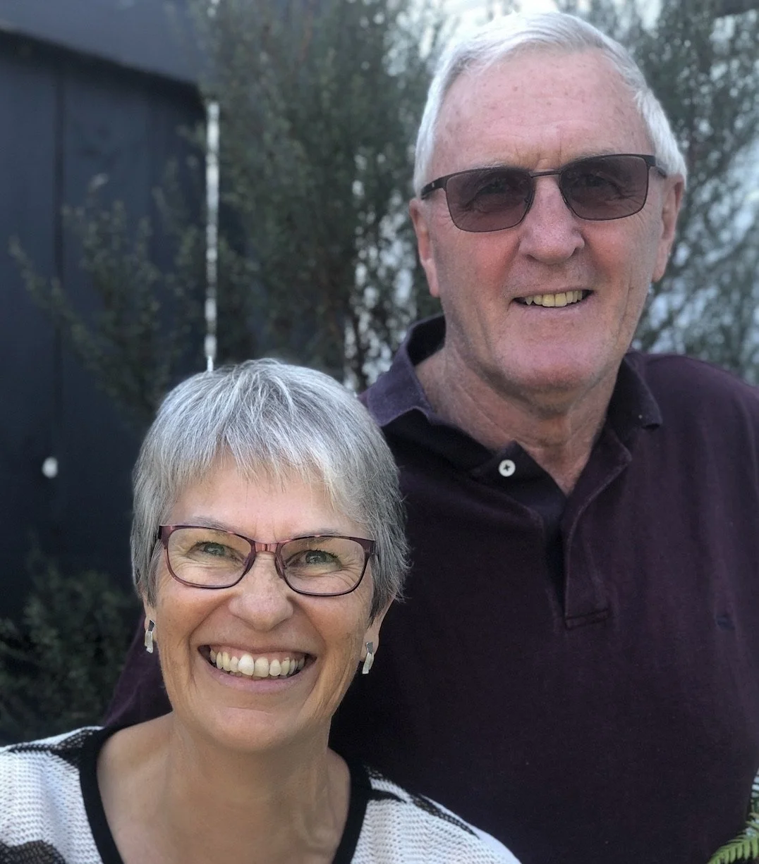 A smiling elderly couple, a woman with short gray hair and glasses and a man with short white hair and sunglasses, standing outdoors with trees in the background.