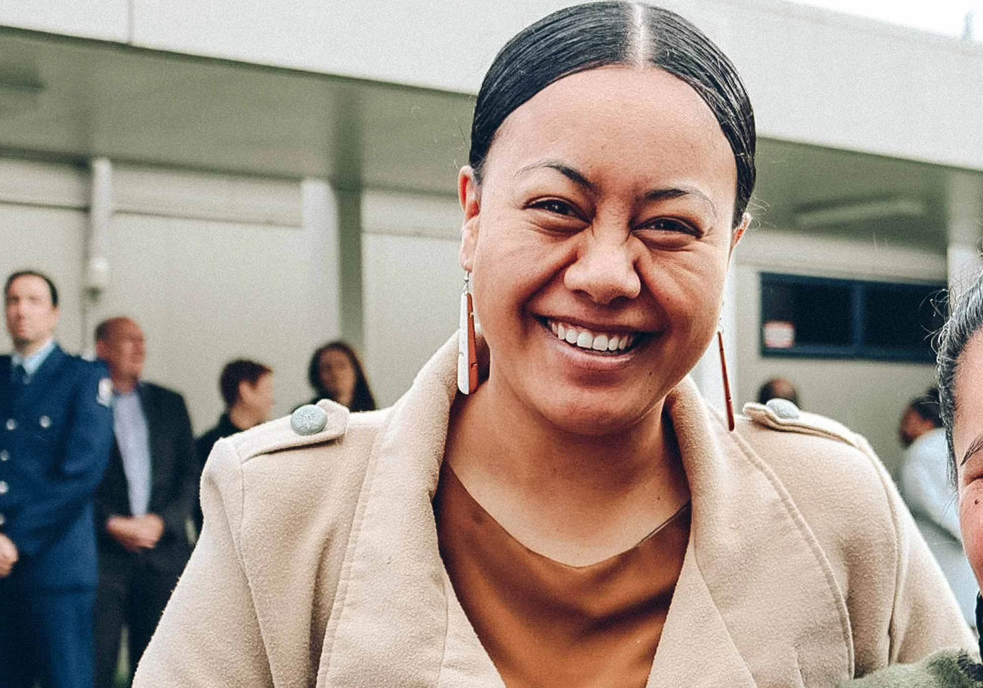 Close-up of a smiling woman with dark hair pulled back, wearing earrings and a beige coat, in a crowded indoor setting.