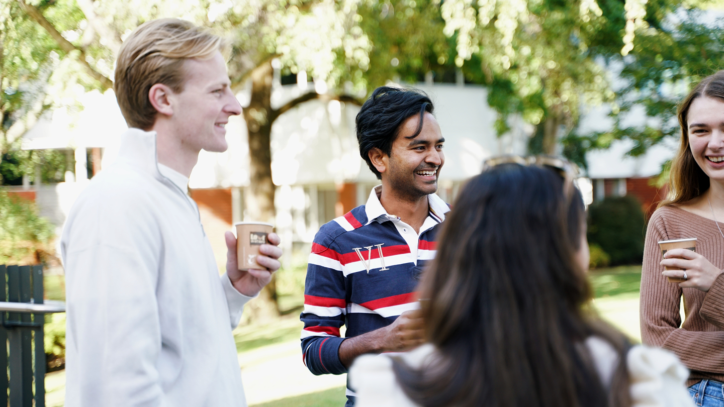 Group of four young adults socializing outdoors, smiling, and holding coffee cups in a park or garden setting.