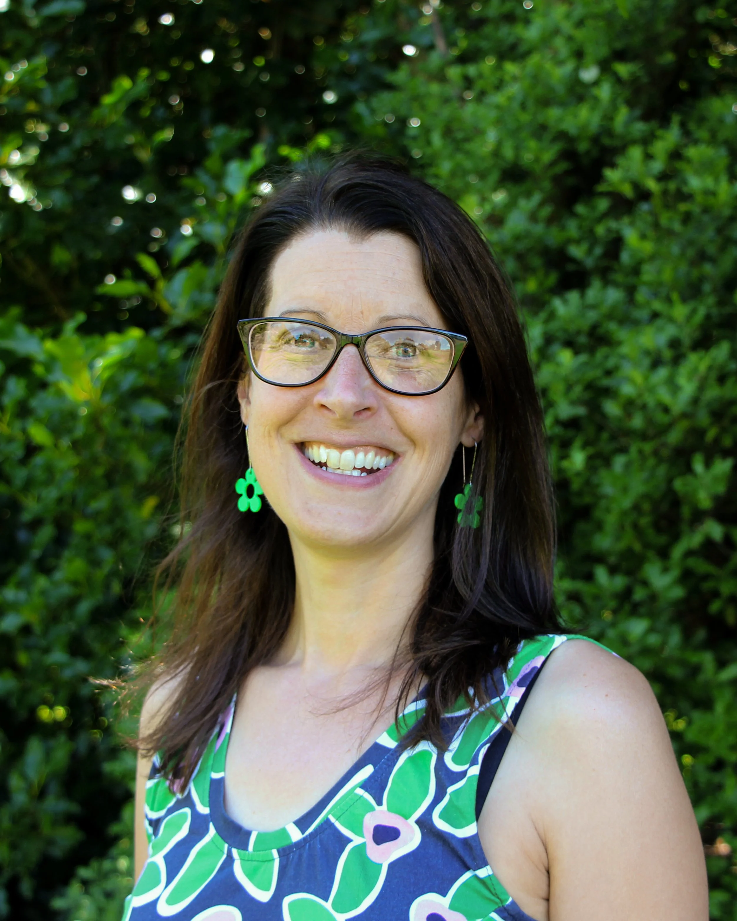 A woman with brown hair wearing glasses and green flower-shaped earrings, smiling outdoors with a green leafy background.