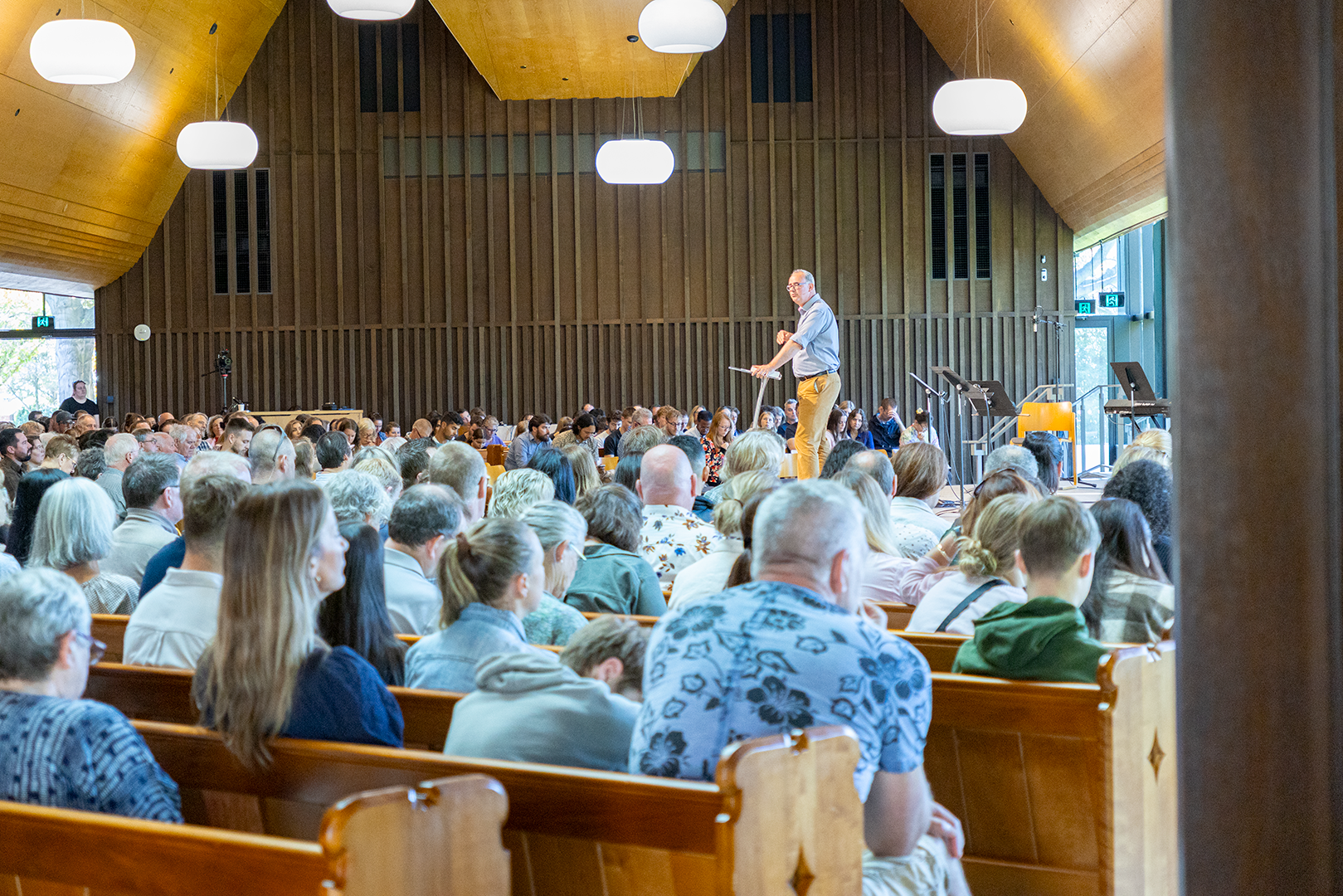 A large gathering of people seated in an auditorium listening to a speaker on stage. The auditorium has wooden walls and ceiling, with hanging white lights. The speaker, a man, stands at a podium facing the audience, with music stands and chairs on stage.