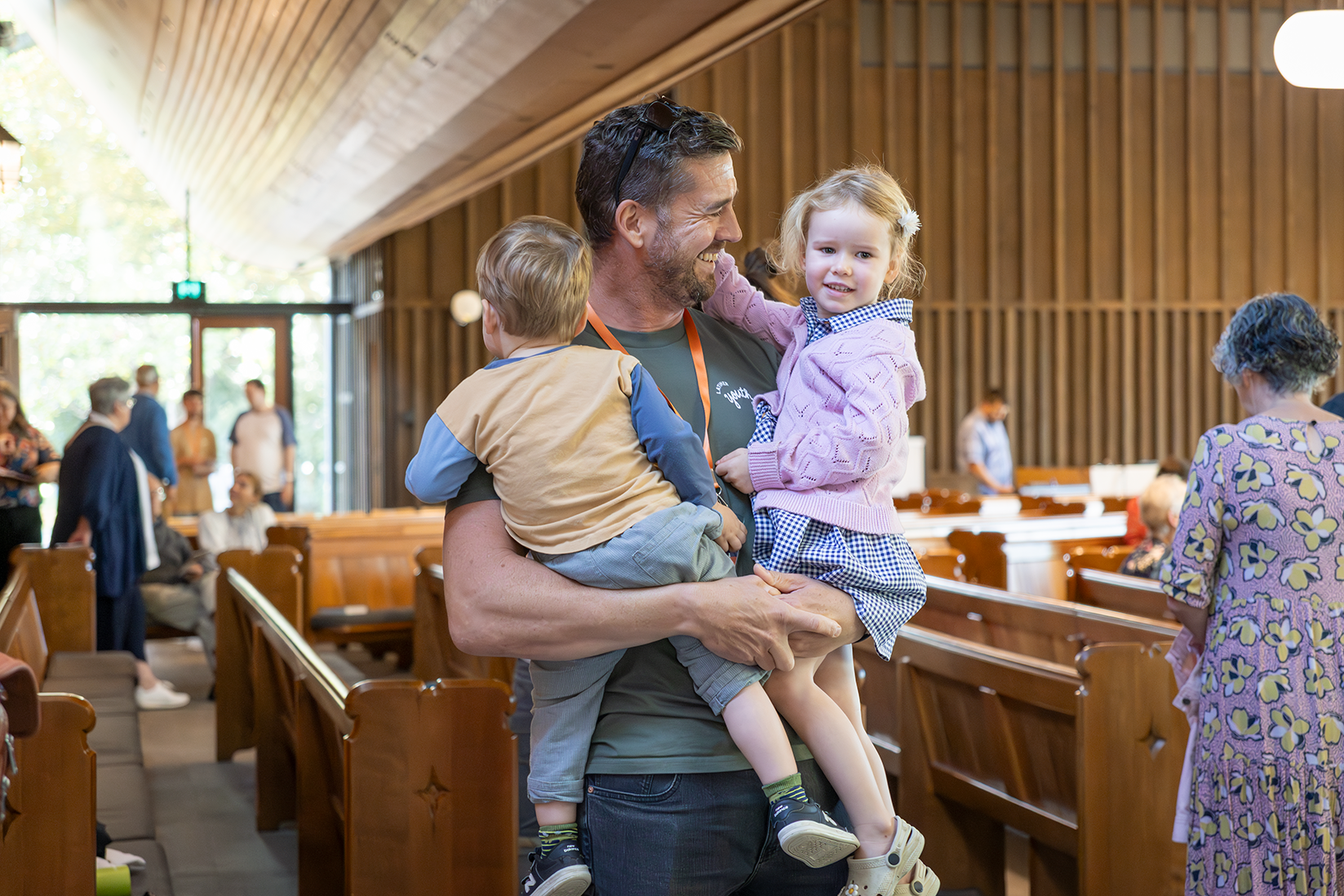 A man holding two children inside a wooden church or chapel, with other people in the background.