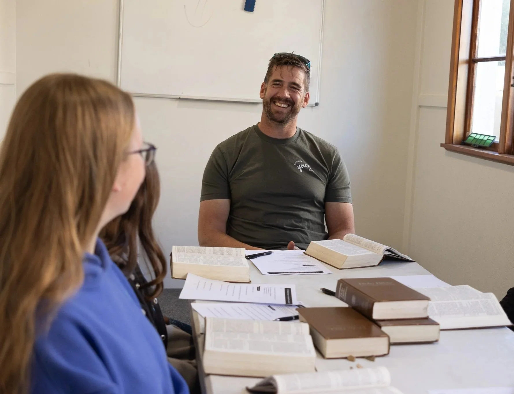 A man and a woman are sitting at a table in a room, with books and papers in front of them. The man is smiling and looking at the woman, who has red hair and is wearing glasses. There is a whiteboard and a window in the background.