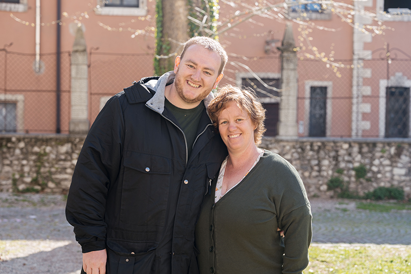 A young man and an older woman smiling outdoors in front of a stone wall and a pink building, with trees and sunlight.