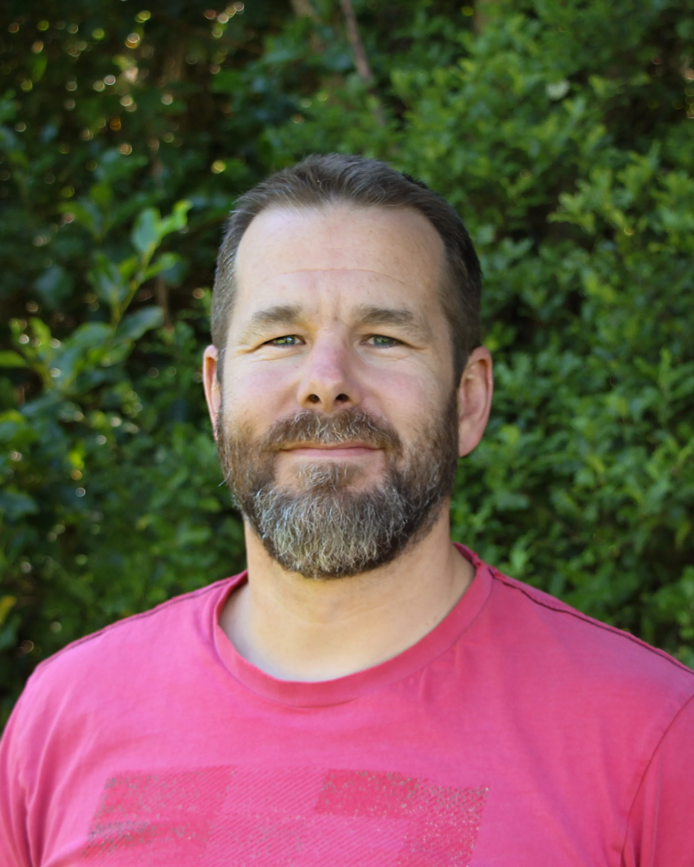 Close-up of a smiling man with a beard and short dark hair, wearing a pink shirt, outdoors with green foliage in the background.