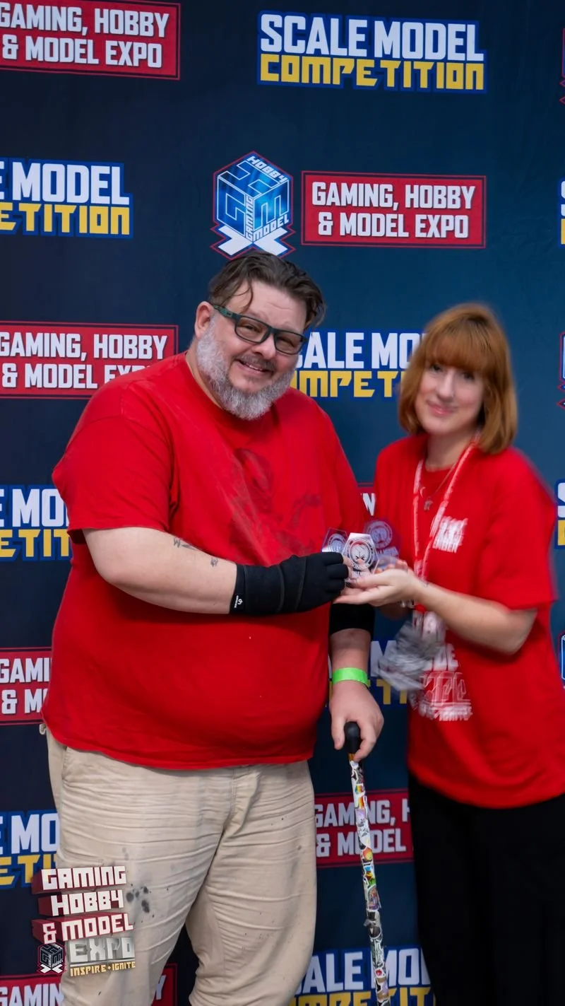 Two people at a gaming, hobby, and model expo standing in front of a backdrop with logos. The man is wearing glasses, a red shirt, beige pants, and gloves, holding a cane and a small clear plastic case. The woman has red hair, is wearing a red shirt and lanyard, and is smiling while handing the man an award or certificate.