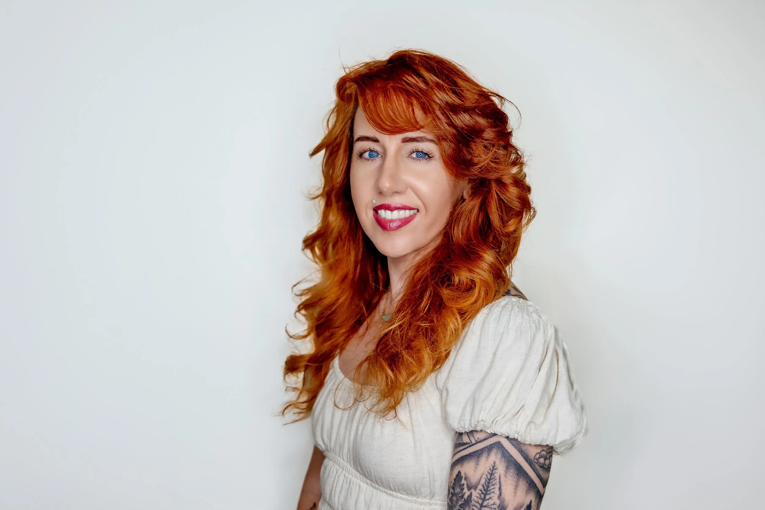 A woman with long, curly red hair and blue eyes, smiling and showing her teeth, wearing a white blouse with puffy sleeves and a tattoo on her arm, standing against a plain light-colored background.
