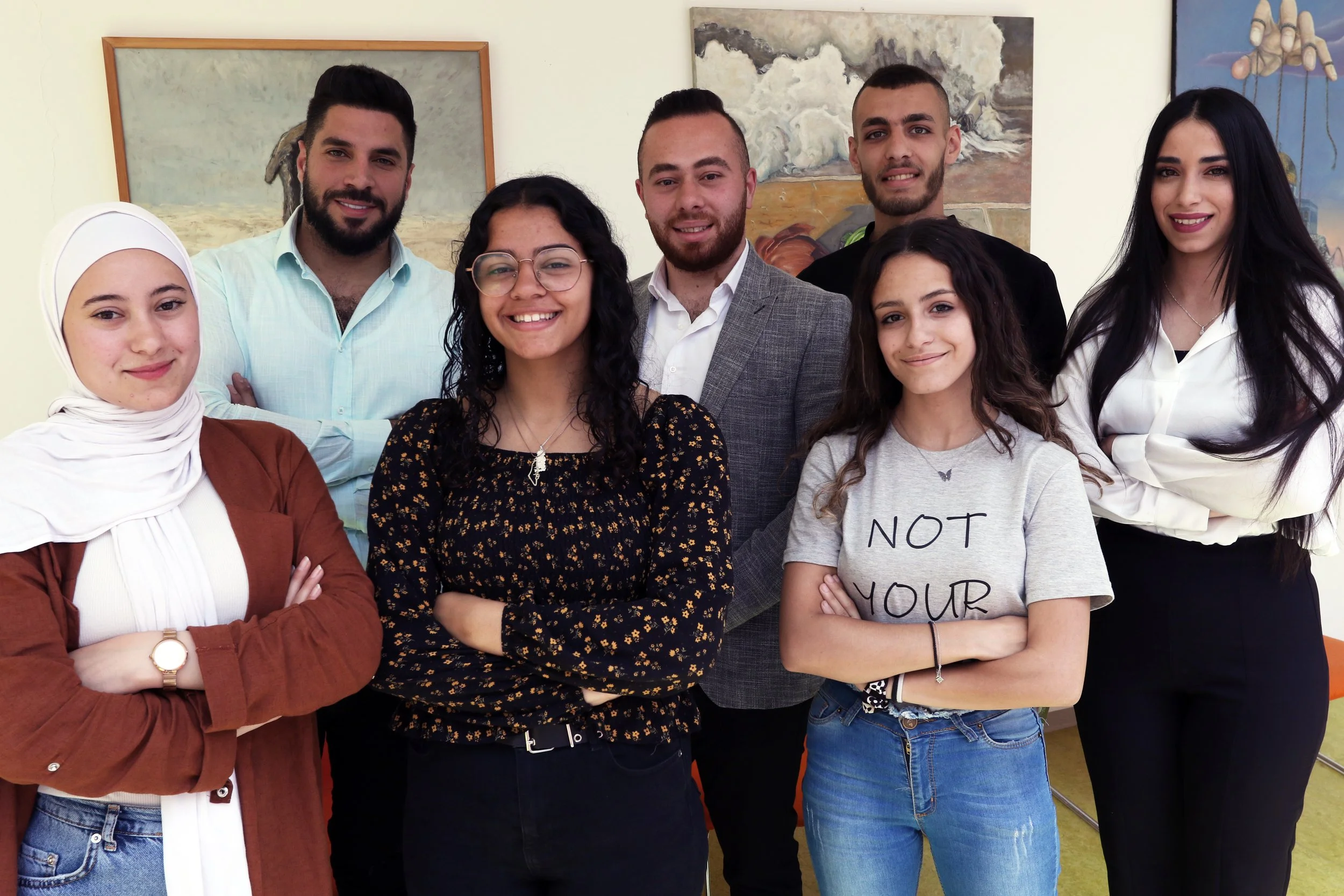 Group of eight young adults, diverse in ethnicity, posing together indoors with paintings on the wall behind them.
