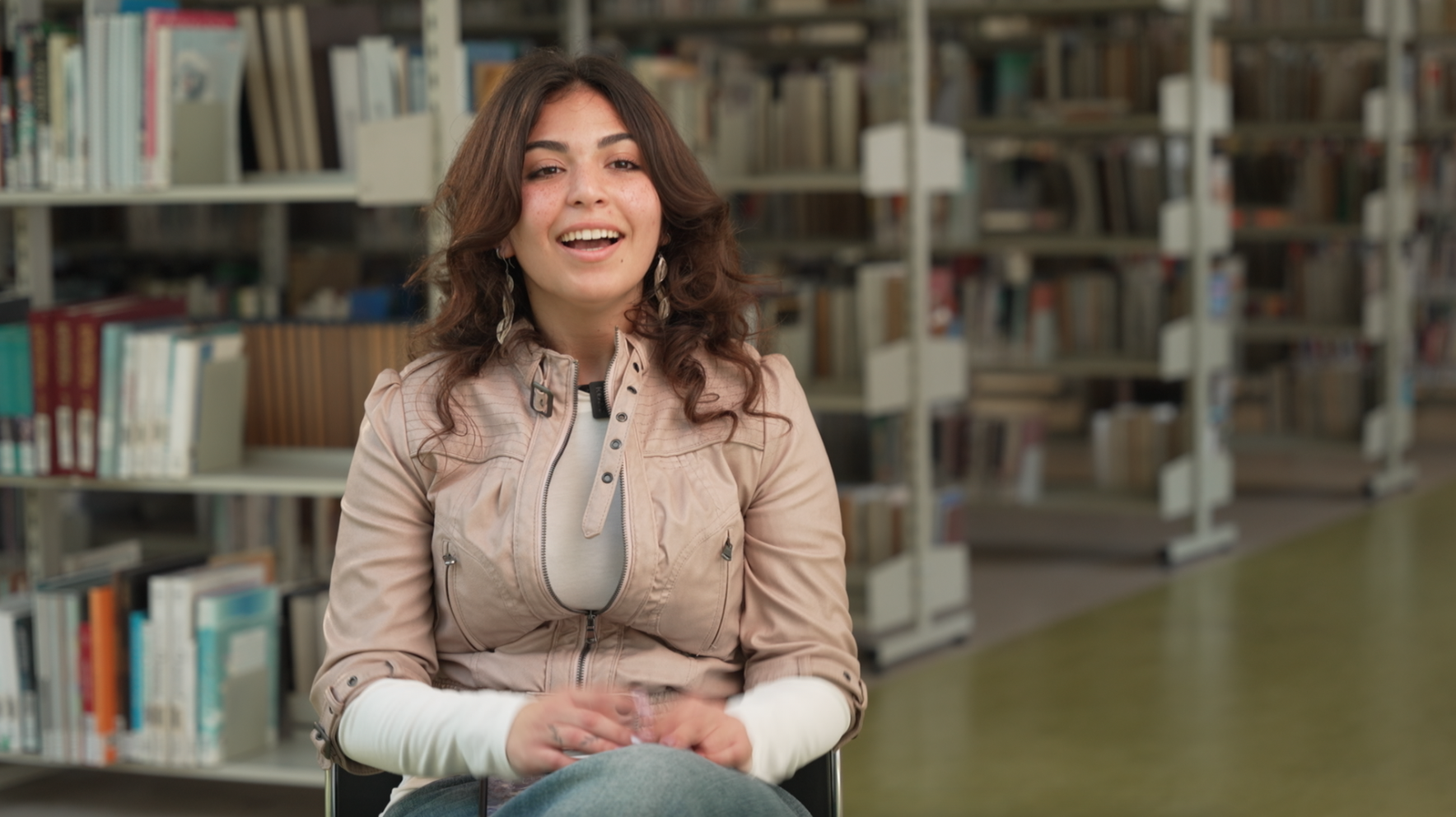 A young woman with curly brown hair, wearing a beige jacket and earrings, sitting in a library aisle, smiling at the camera.