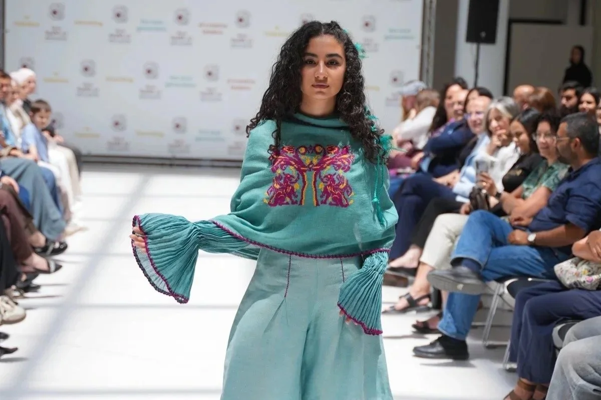 Female model walking on the runway at a fashion show, wearing a green sweater with colorful animal design and pleated bell sleeves, with an audience watching.