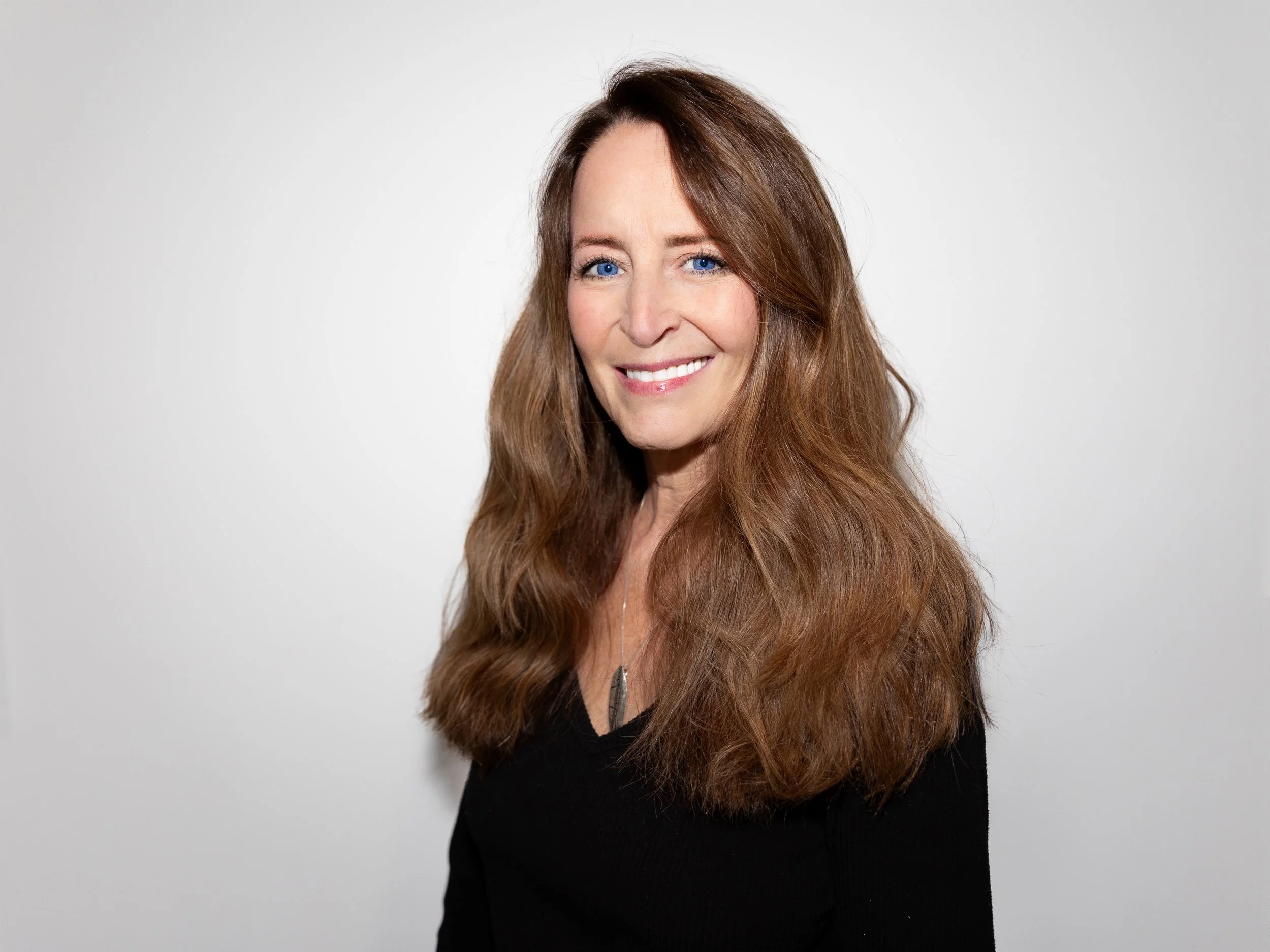 Portrait of a woman with long, wavy brown hair, blue eyes, and a bright smile, wearing a black top and standing against a plain white background.
