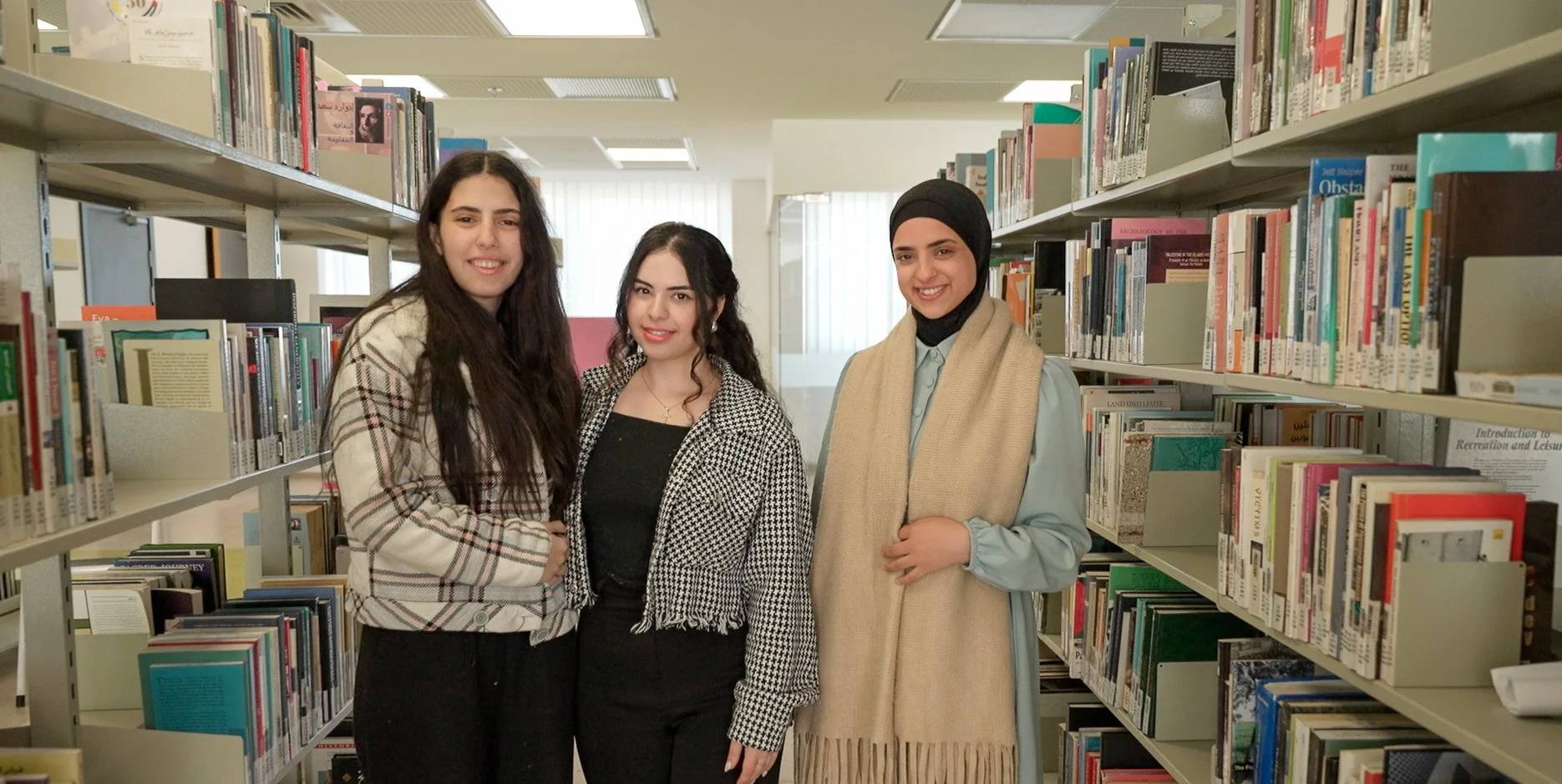 Three young women standing together in a library aisle between bookshelves filled with books, smiling at the camera.