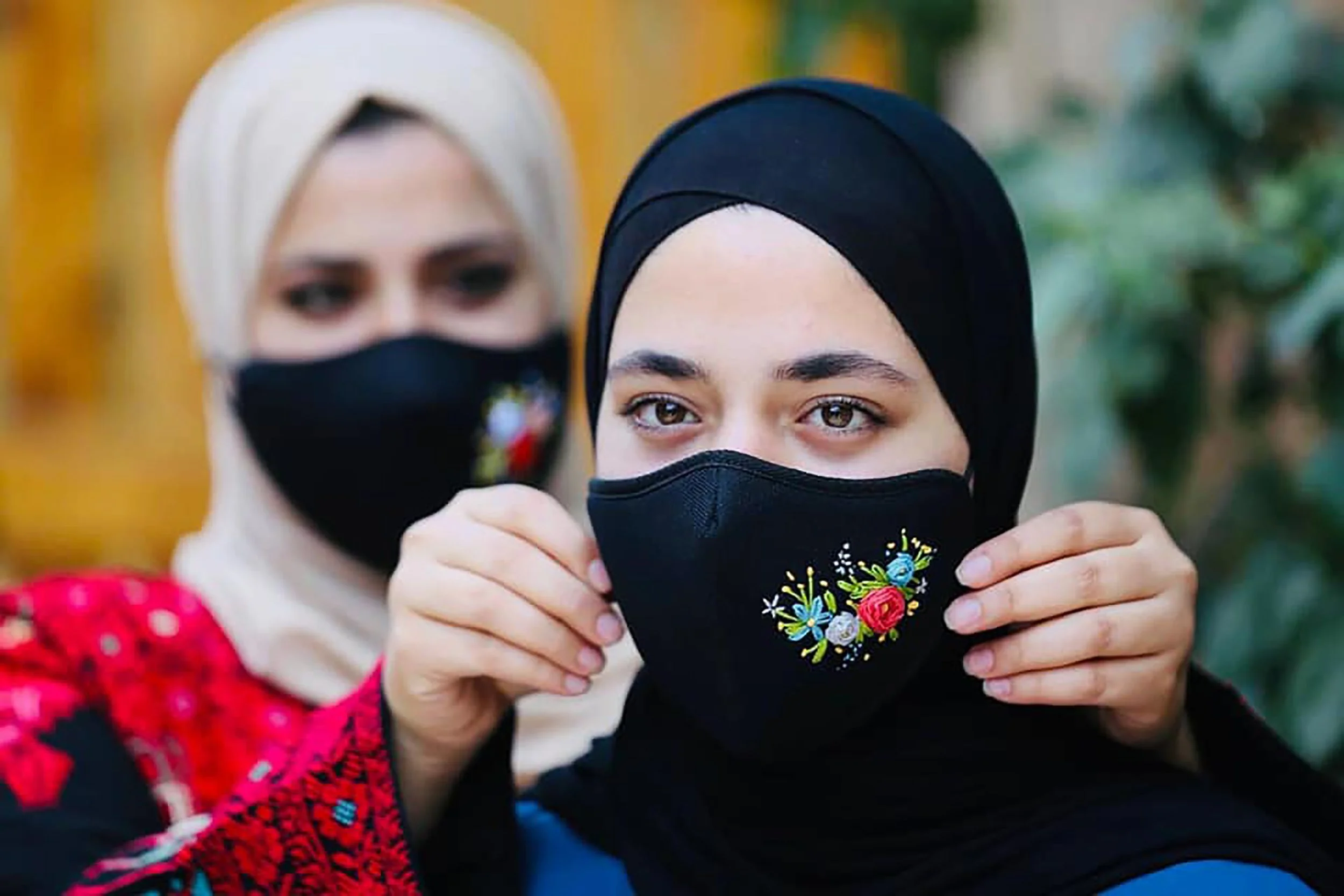 A woman with brown eyes and wearing a black hijab holds a black face mask with colorful floral embroidery, while another woman with light skin, brown eyes, wearing a beige hijab, and a red dress with white polka dots stands behind her.
