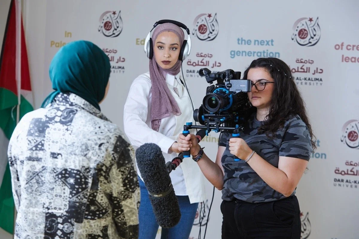 Two women filming an interview at Dar Al-Kalima University, with a logo and text in the background. One woman wears a blue hijab and patterned jacket, the other wears glasses and a camouflage shirt, operating a professional camera. The woman being filmed is visible from behind.