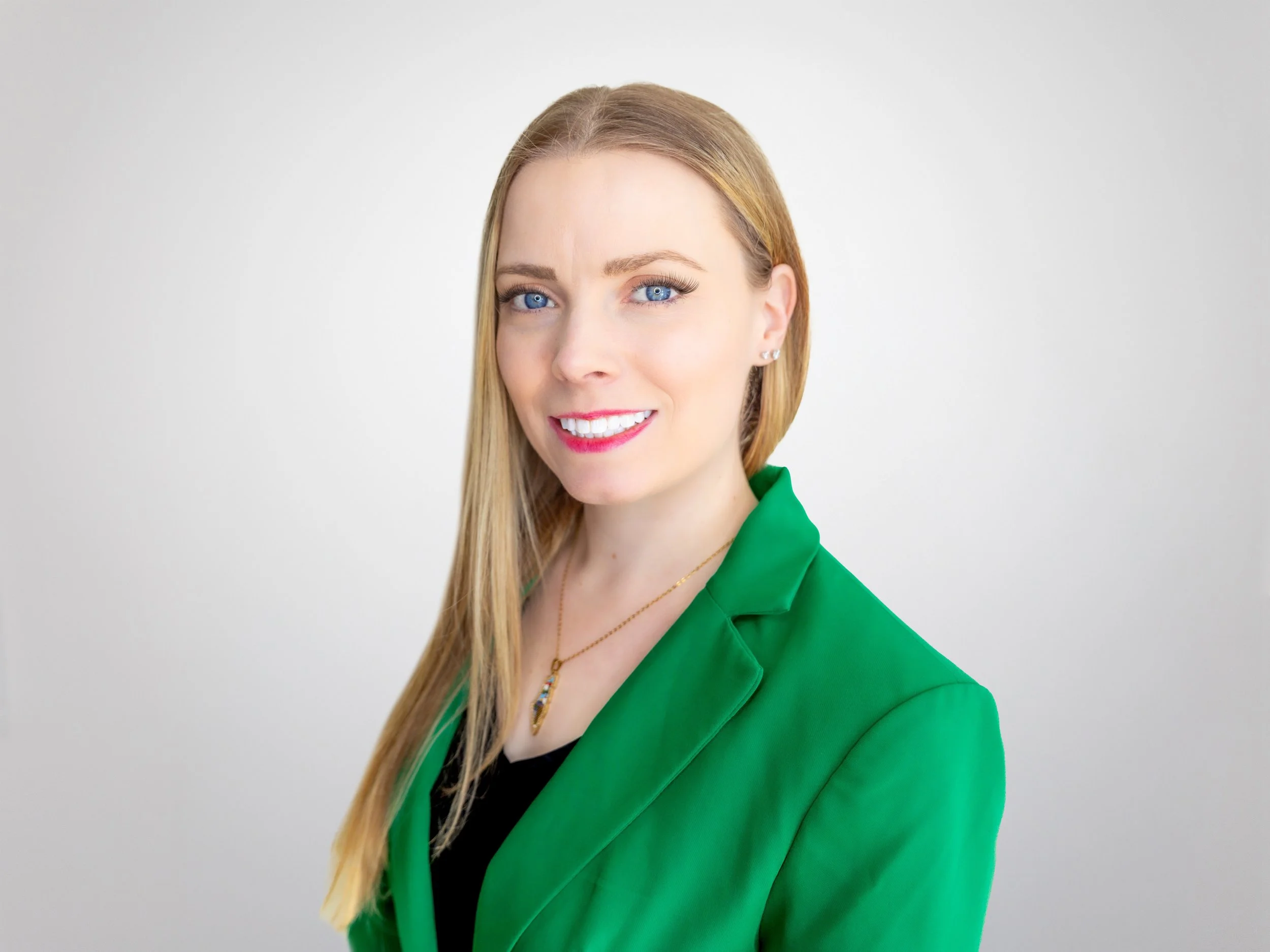A professional woman with long blonde hair, wearing a green blazer, smiling, and standing against a plain white background.