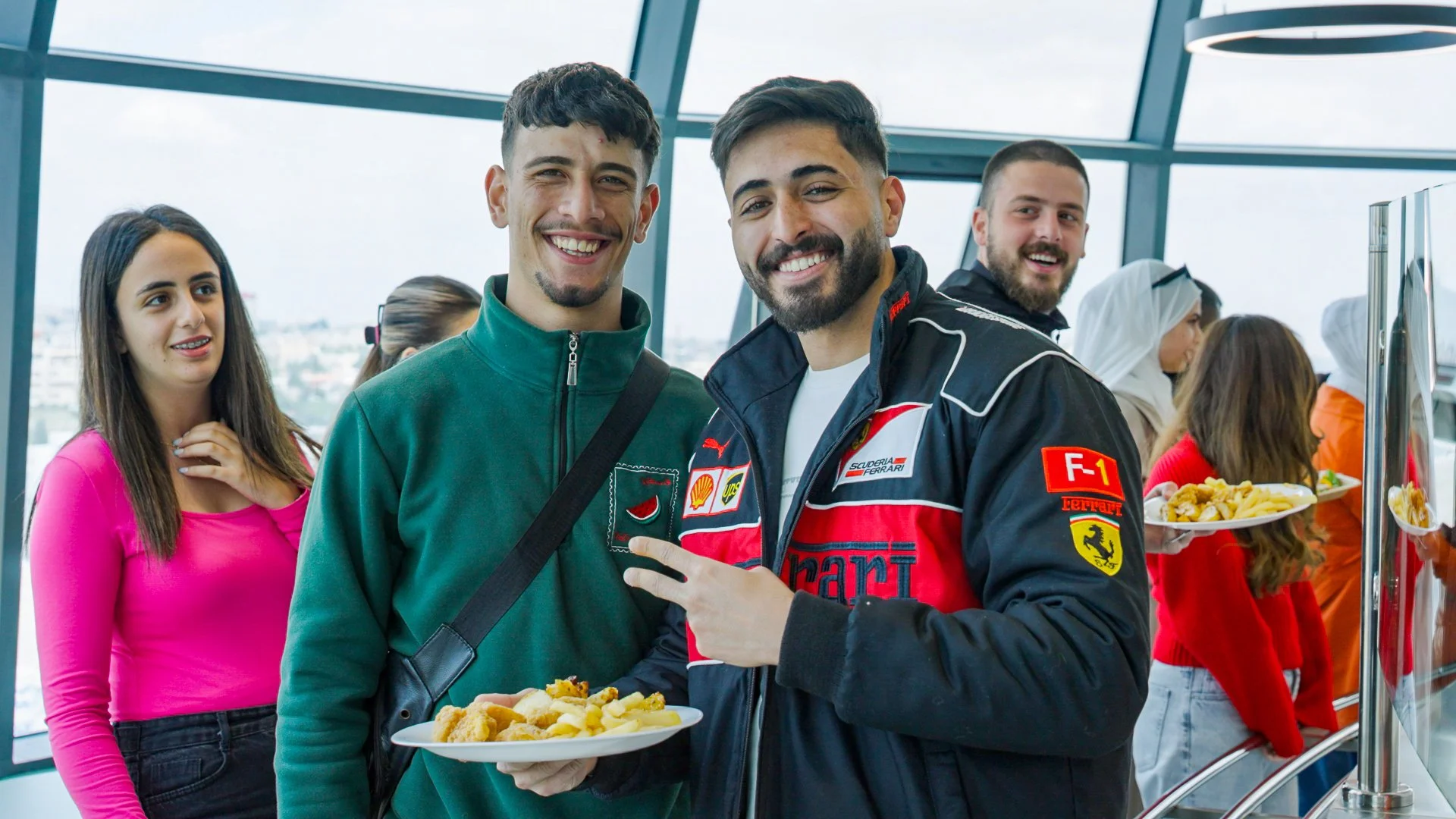 Smiling group of people at a buffet, some holding plates of food, in a bright, modern space with large windows.