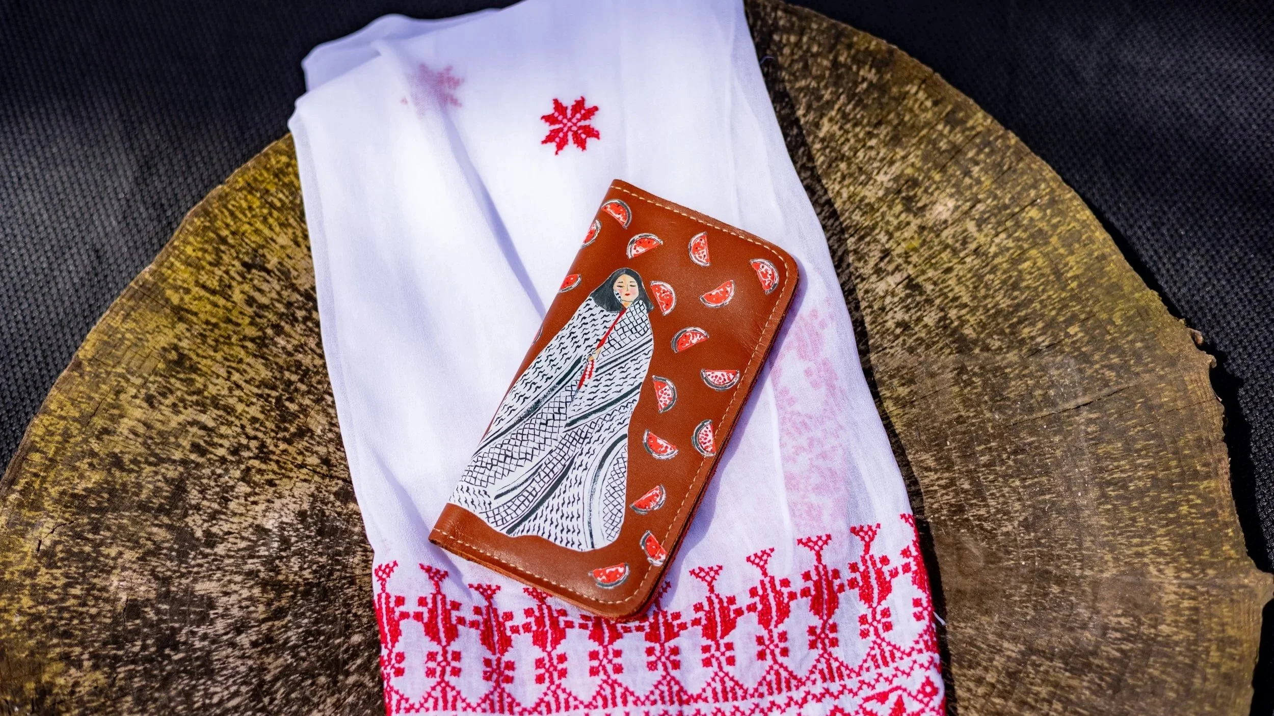A white cloth with red embroidery, a brown wallet with a woman in traditional attire illustration and red-patterned embroidery, and a wooden surface underneath.