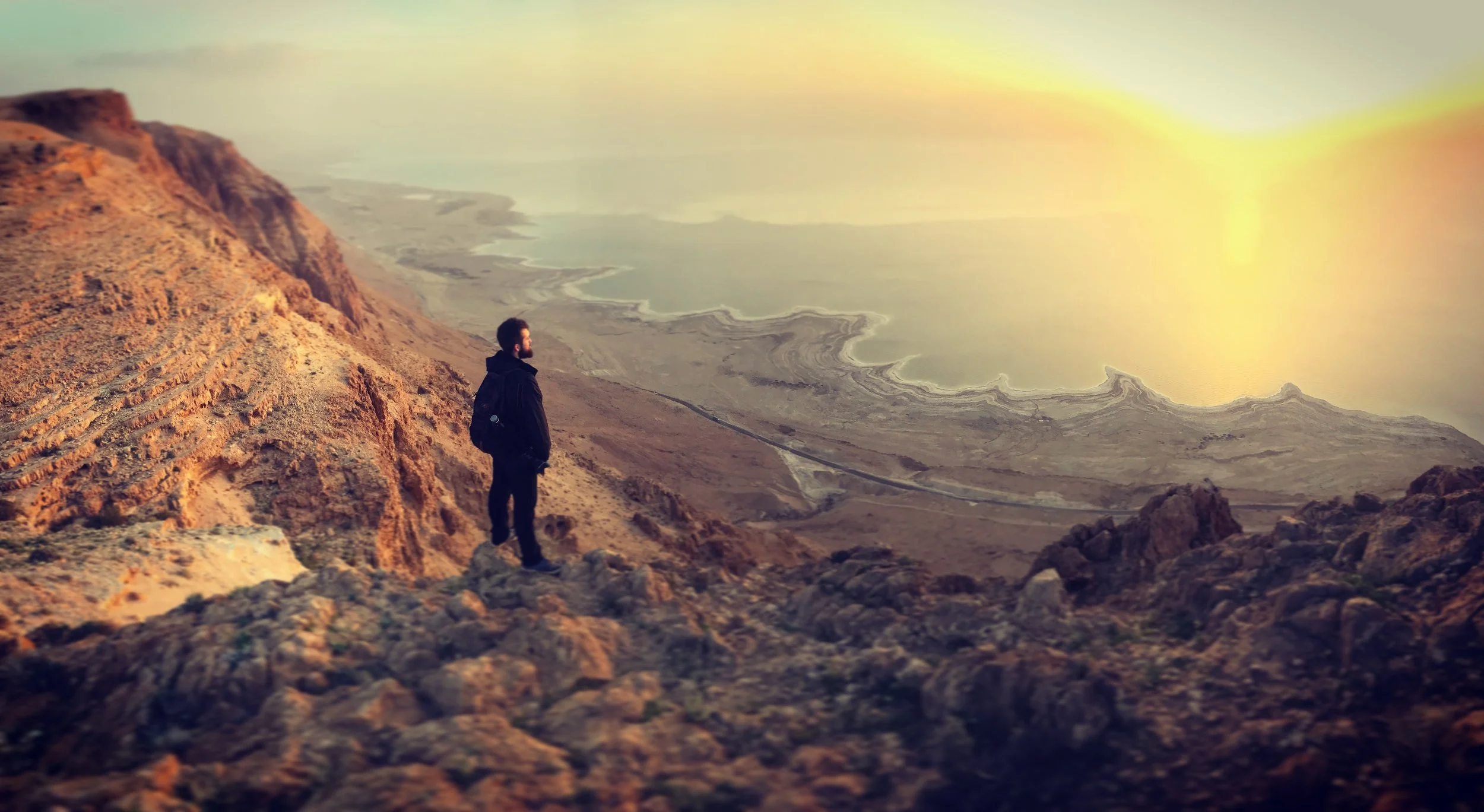 A person standing on rocky terrain overlooking a vast desert landscape and a salt flat, with a sunrise or sunset in the sky.