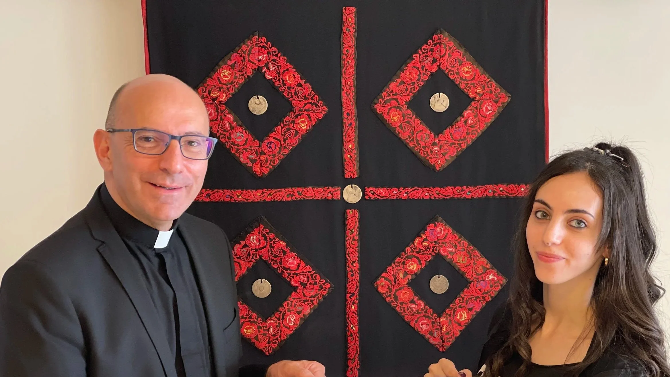 A man in a black clerical shirt and glasses and a woman in a black dress stand in front of a red and black textile wall hanging with square patterns decorated with floral designs and coins, possibly an altar or ceremonial art piece.