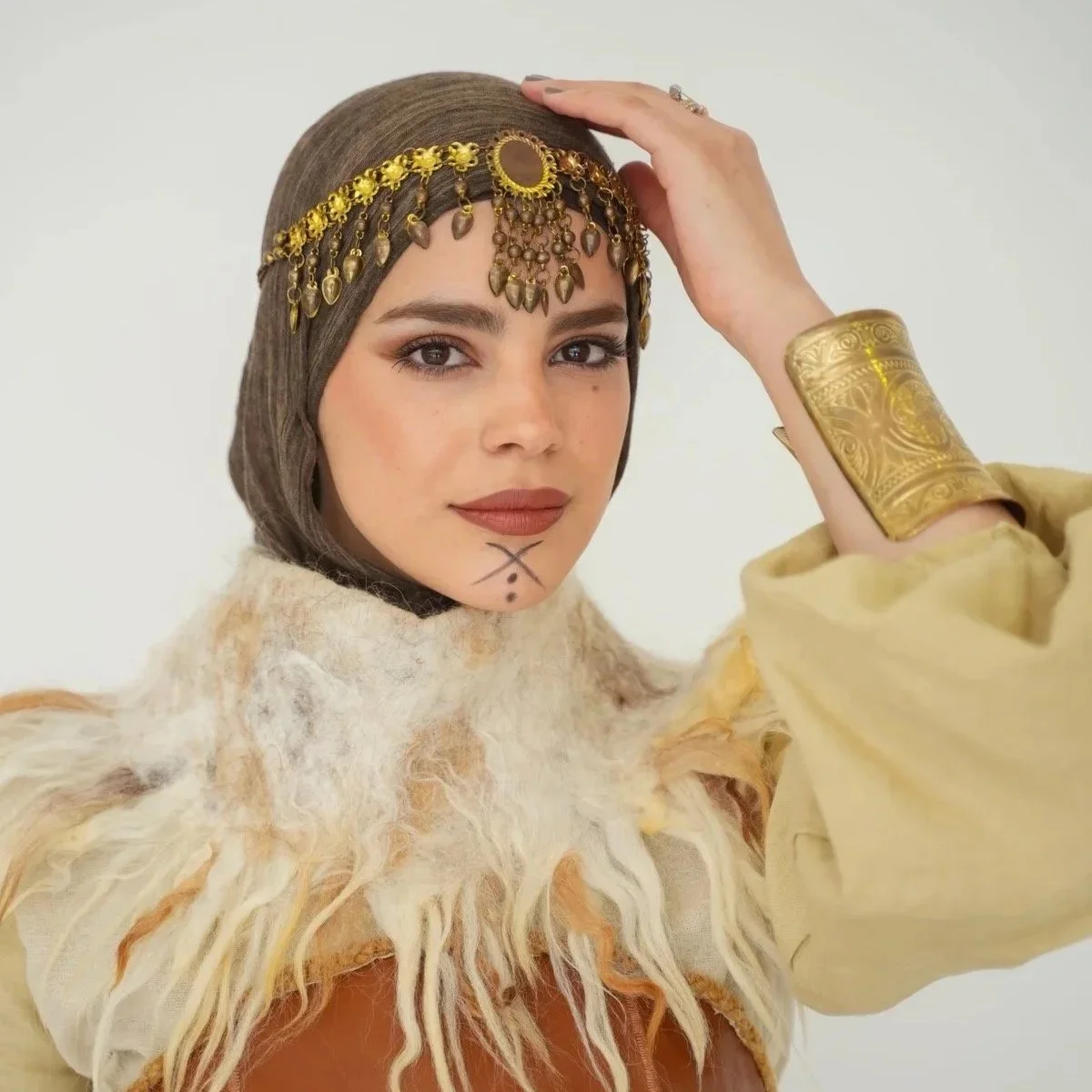Woman wearing a gold headpiece with beads and coins, a large gold bracelet, and a beige and brown fur-like garment, with face markings.