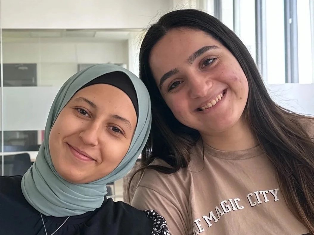 Two young women smiling and close together, one wearing a hijab and the other with long dark hair, in an indoor setting with windows in the background.