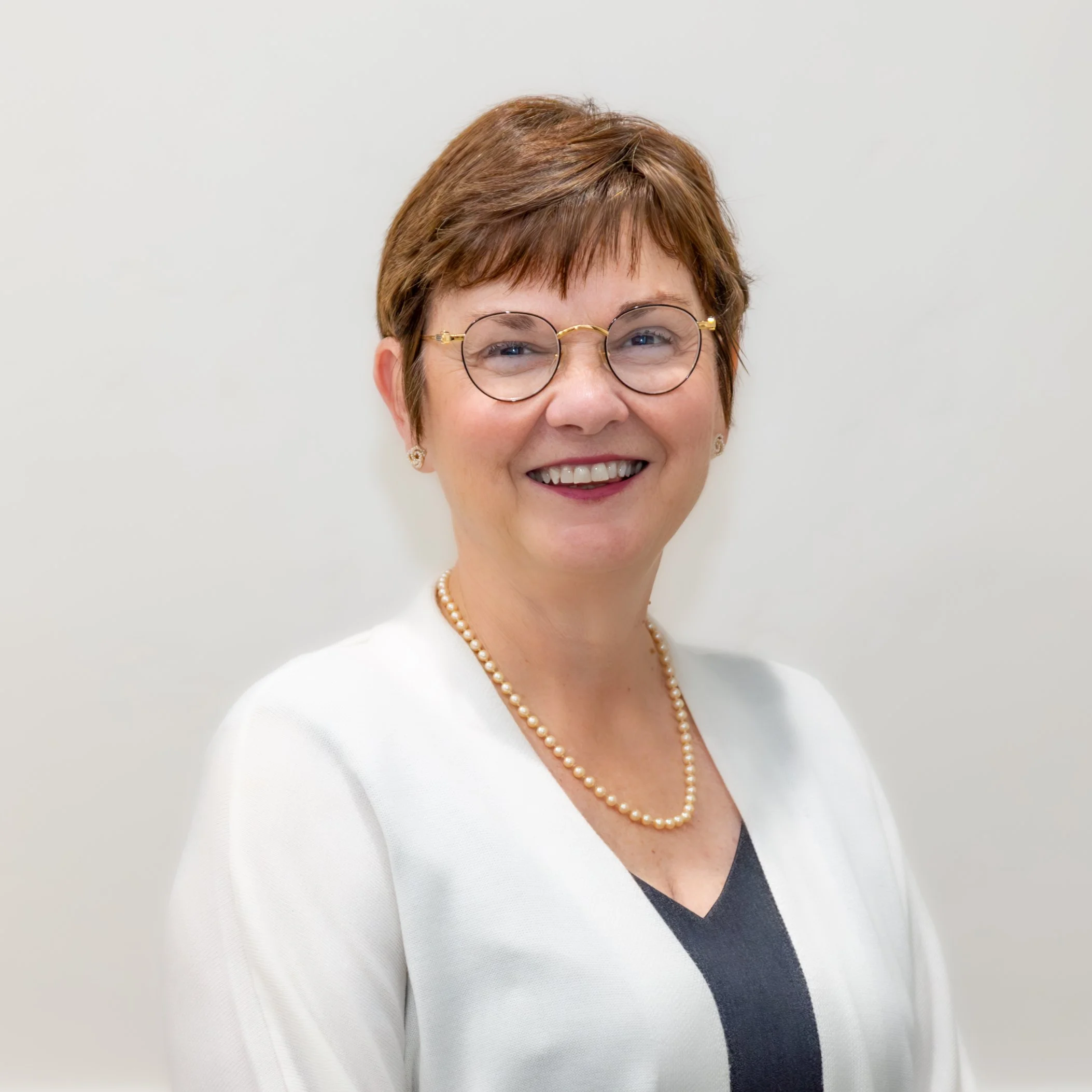 Portrait of a smiling woman with short brown hair, glasses, pearl necklace, and white blazer against a plain light background.