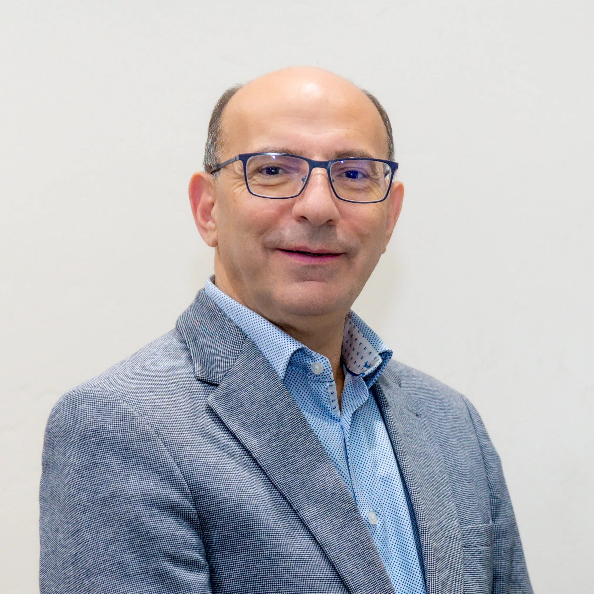A man wearing glasses and a light gray blazer, standing against a plain white wall.