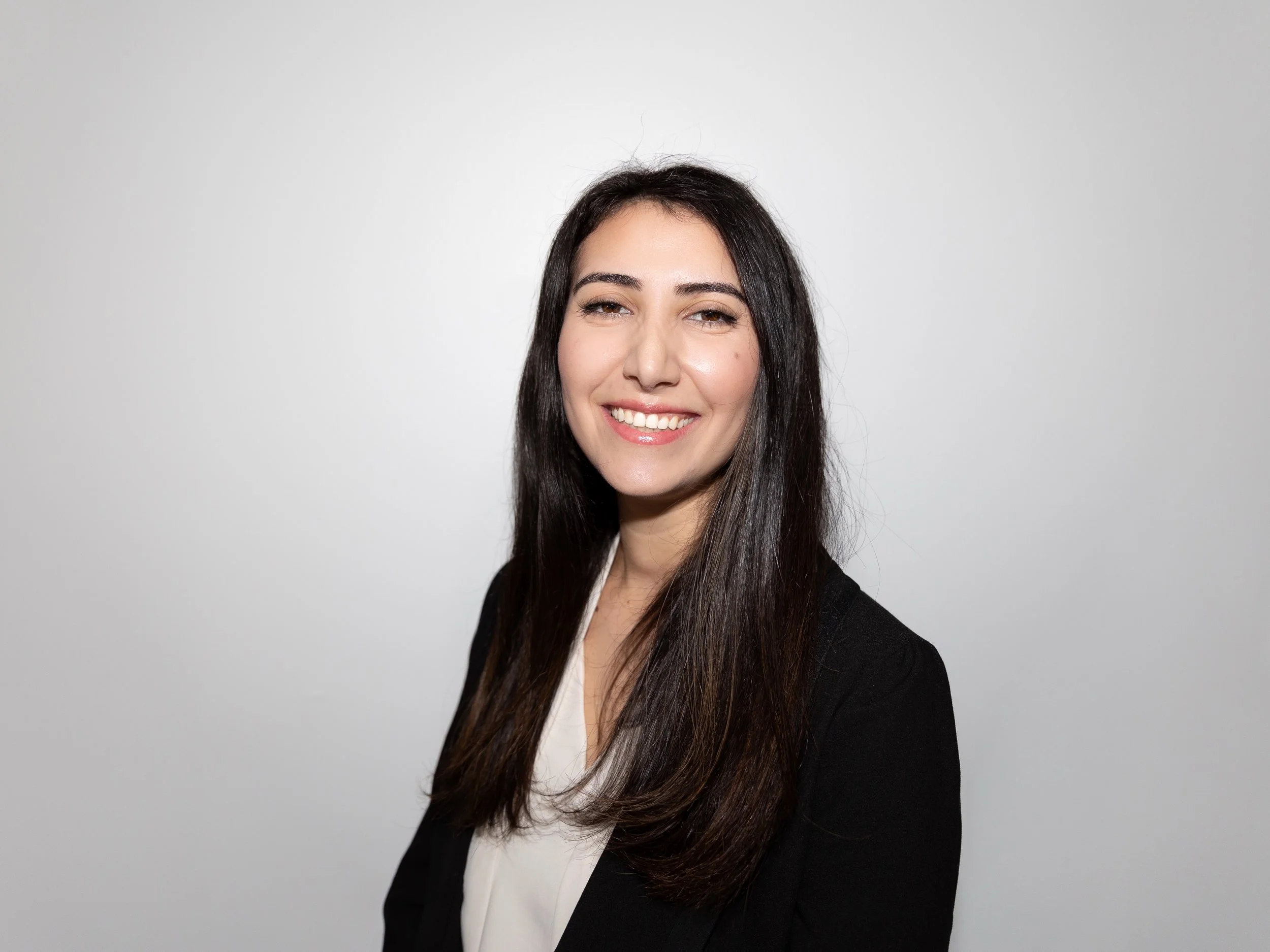 A woman with long dark hair, wearing a black blazer and white top, smiling against a plain light gray background.