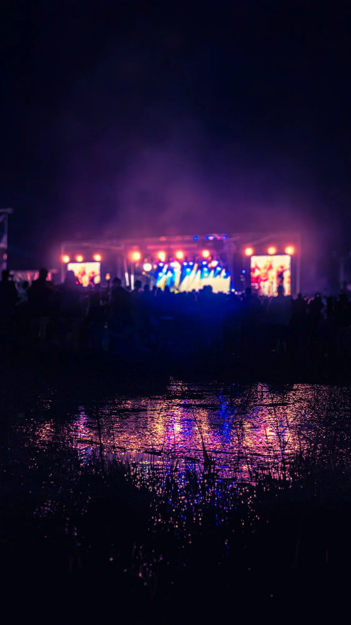 Nighttime outdoor concert stage with bright colorful lights, reflected on a body of water with silhouettes of people watching.