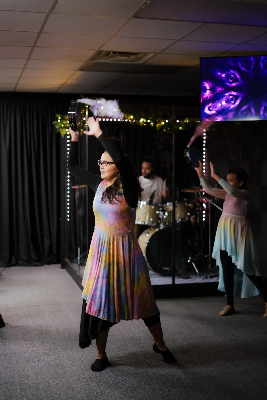 Woman in a colorful dress with black long sleeves dancing and holding a tambourine during a performance live music event with band and decorated stage.