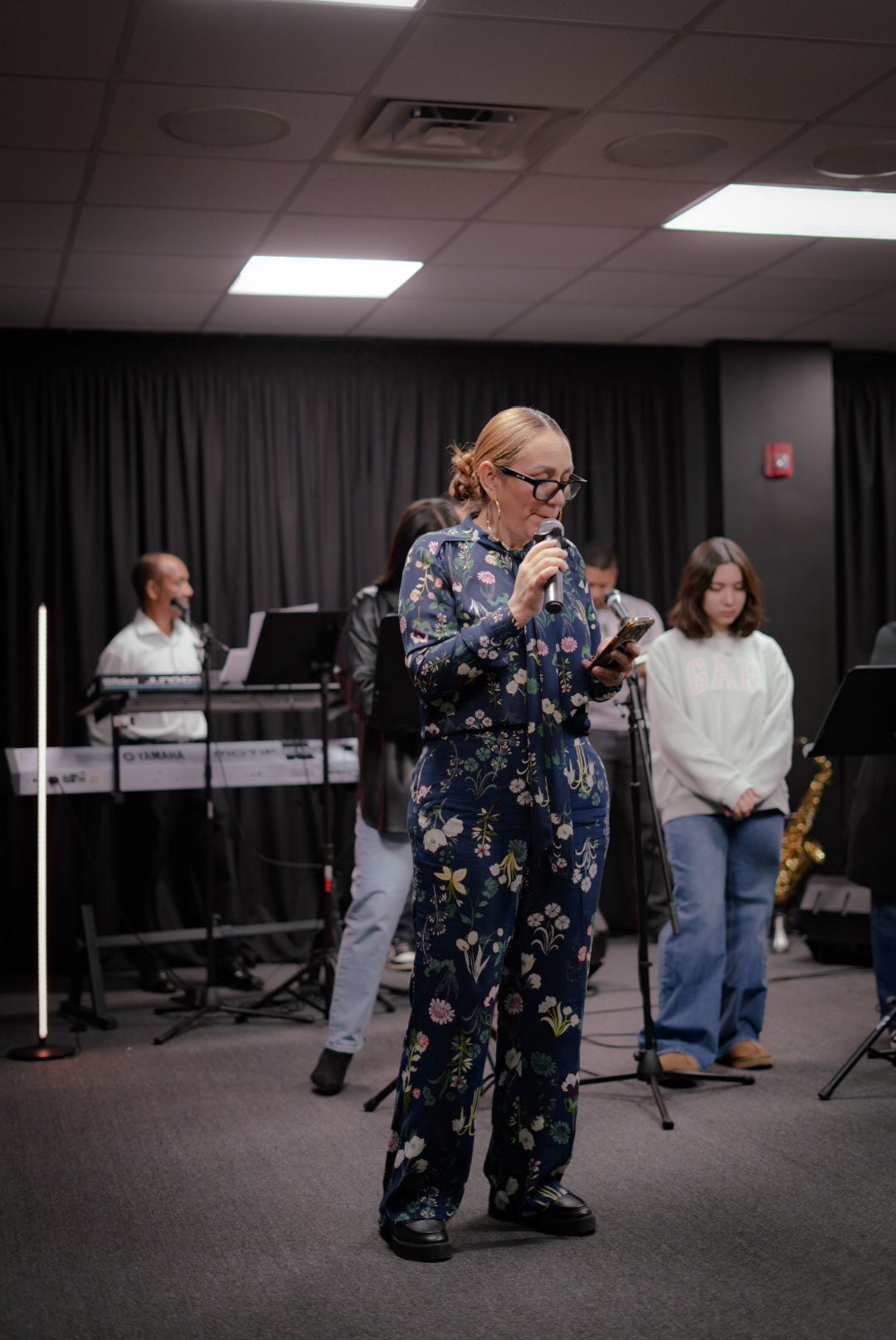 A woman with glasses and a floral jumpsuit speaks into a microphone while reading from her phone, in front of a stage with a band, including a keyboard, and other musicians, in a dimly lit indoor event space.