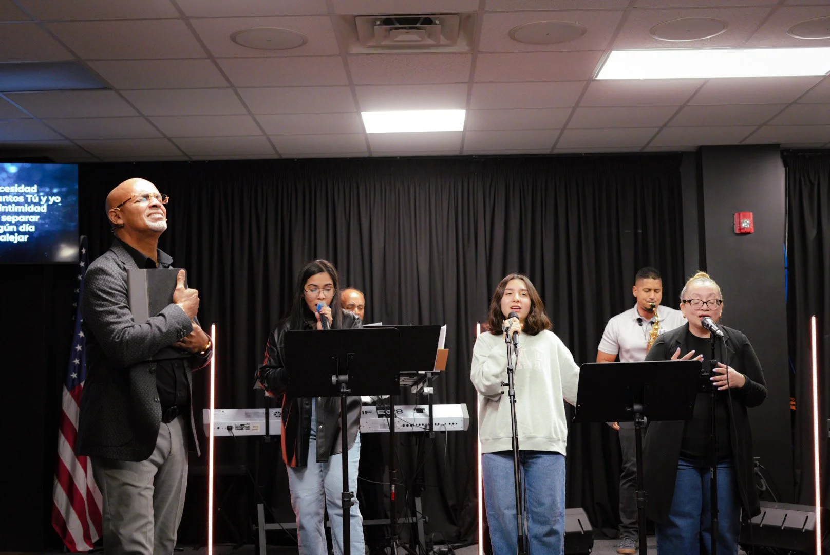 Group of people singing and playing music on stage with a black curtain background, microphones, and musical instruments, with a man holding a folder and several young women singing.