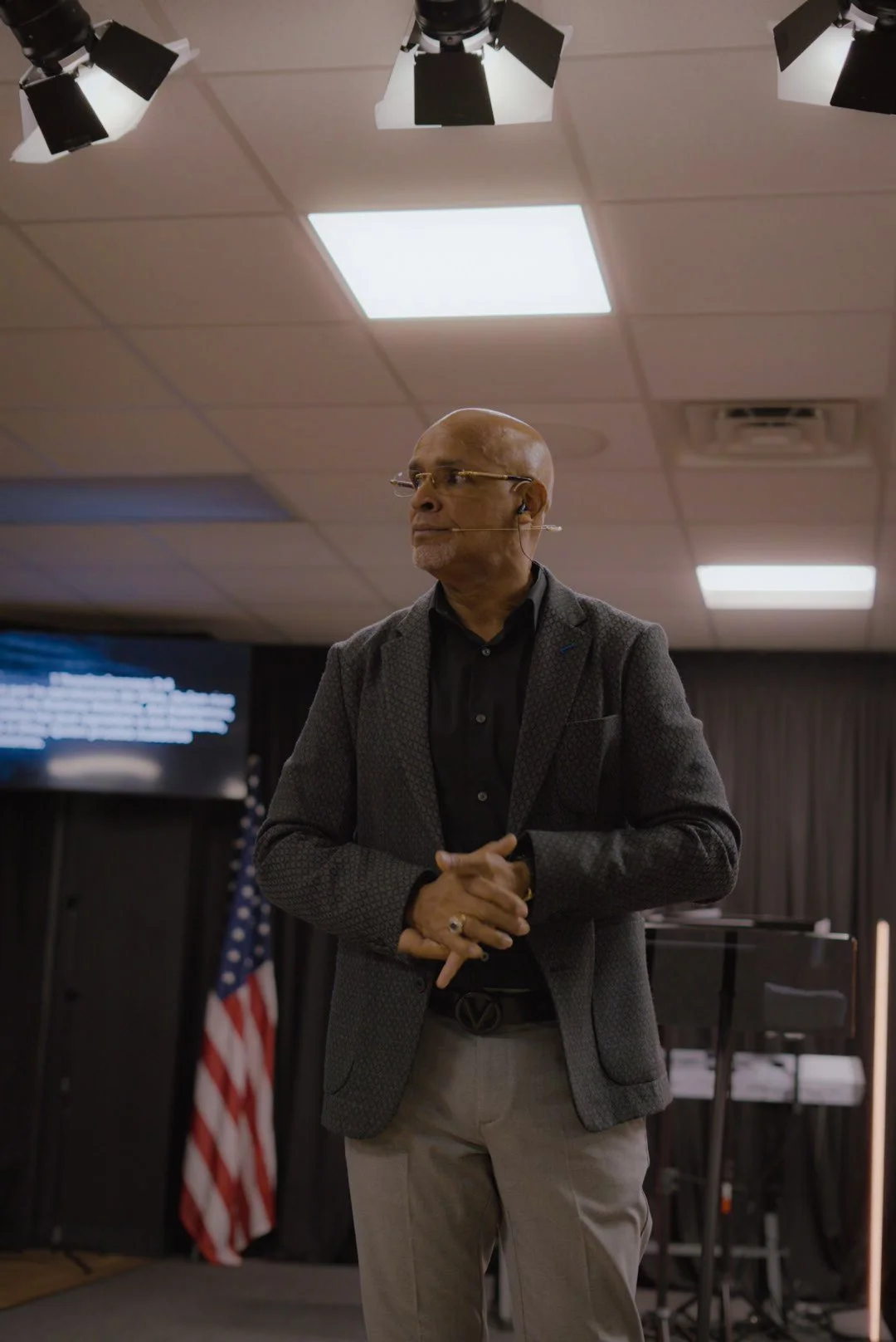 A man wearing glasses, a dark blazer, and a microphone headset, standing with hands clasped in a room with a ceiling-mounted light, American flag, and a screen in the background.