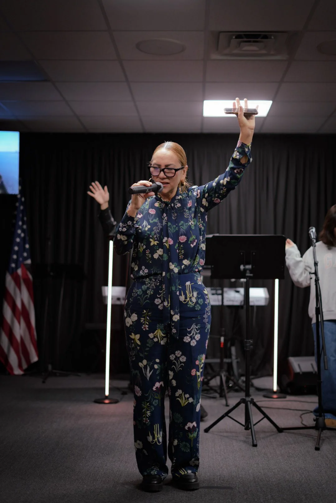 A woman with red hair, glasses, and wearing a floral jumpsuit holding a microphone with her eyes closed and raising her hand during a performance or speech on a stage with musical instruments and an American flag in the background.