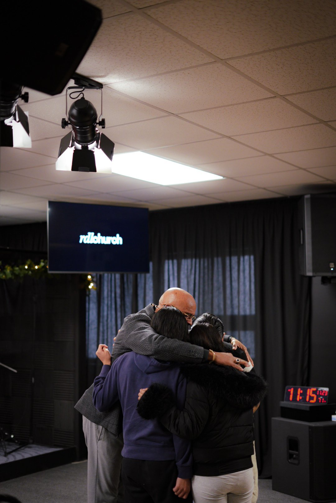 A group of four people embracing in a circle, suggesting a moment of prayer or unity, inside a room with a screen that says 'ridlechurch' and a digital clock displaying 11:15.
