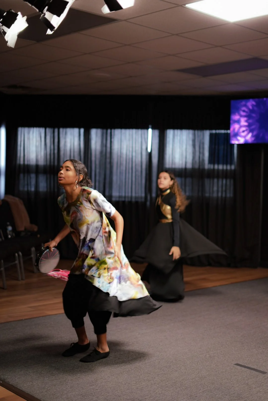 Two young girls performing a dance on stage, one in a colorful dress holding a fan, and the other in a black and gold outfit, with black curtains and a TV screen in the background.
