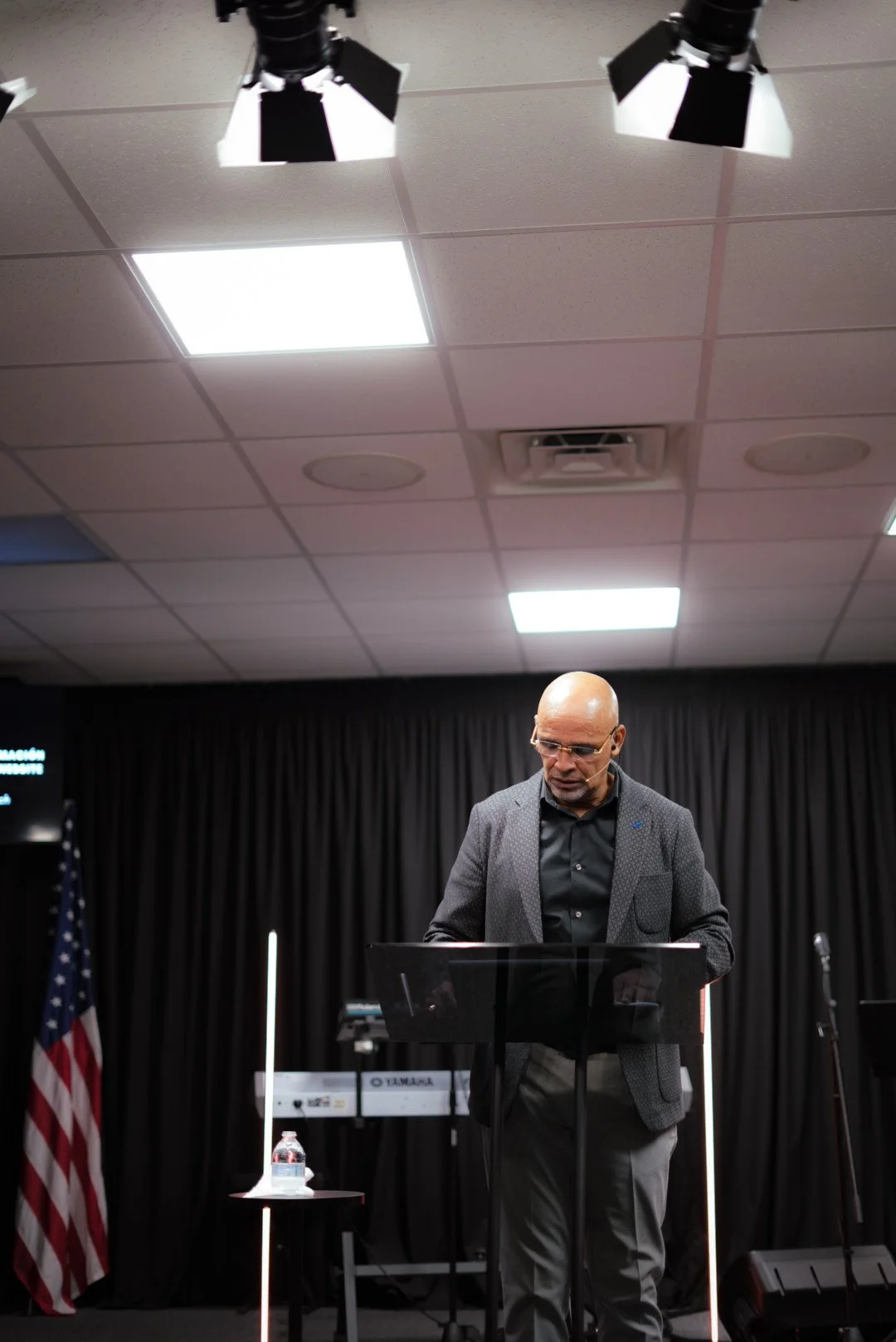 A man with glasses and a bald head stands at a podium in a dark room, with a black curtain behind him, reading or speaking. There is a small table with a water bottle on it, and musical equipment including a keyboard and microphone. The room is lit b
