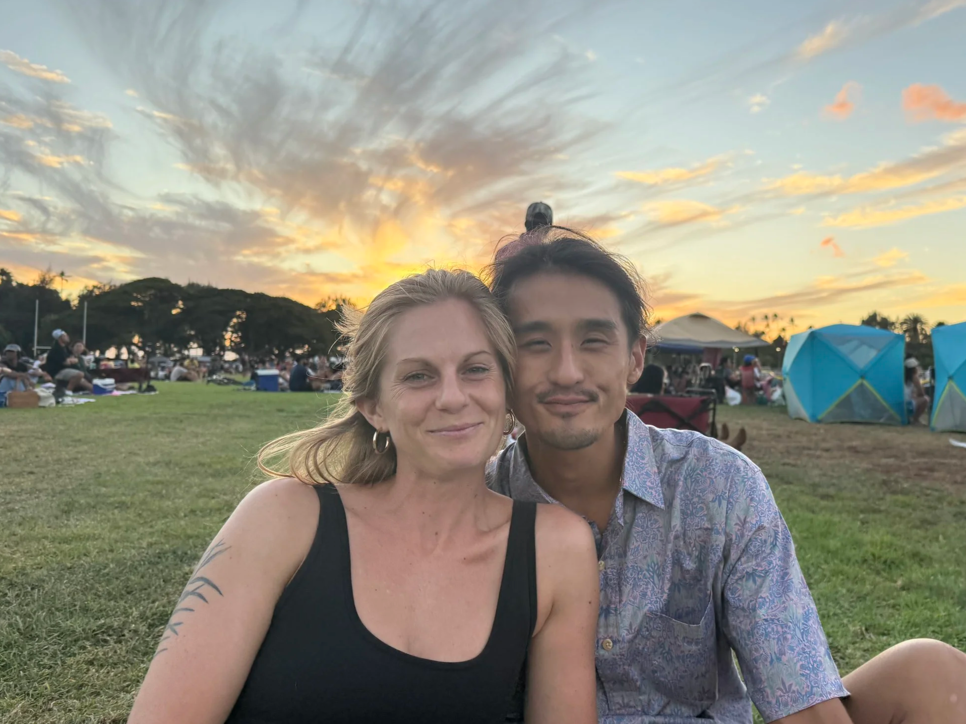 Two people, a woman with blonde hair and a man with dark hair, smiling and sitting close together outdoors during sunset with a sky filled with clouds. In the background, there are people sitting on the grass, tents, and trees.