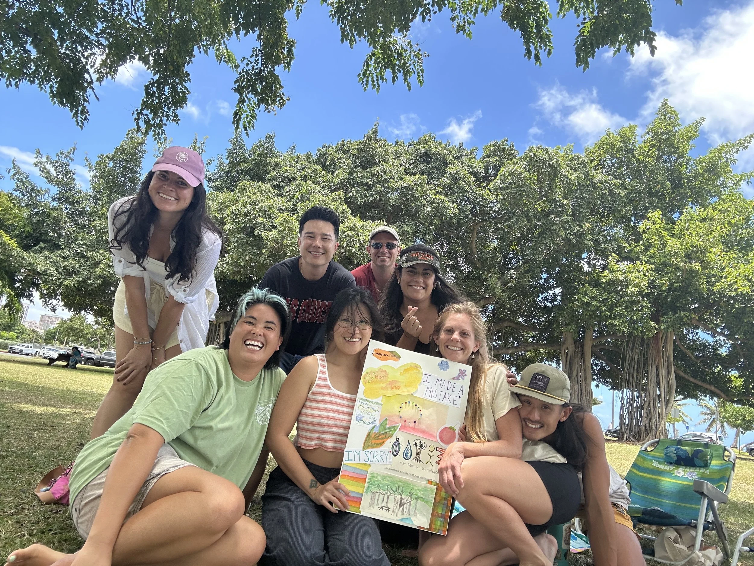 Group of smiling people outdoors beneath trees, holding a colorful poster with drawings and text, sunny sky in background.