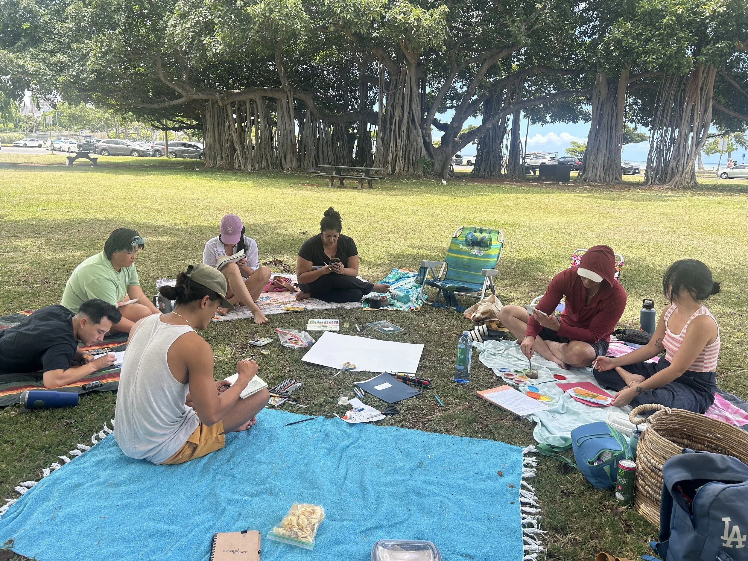 Group of people sitting on blankets and chairs beneath a large tree, engaging in drawing, writing, and chatting in a park.