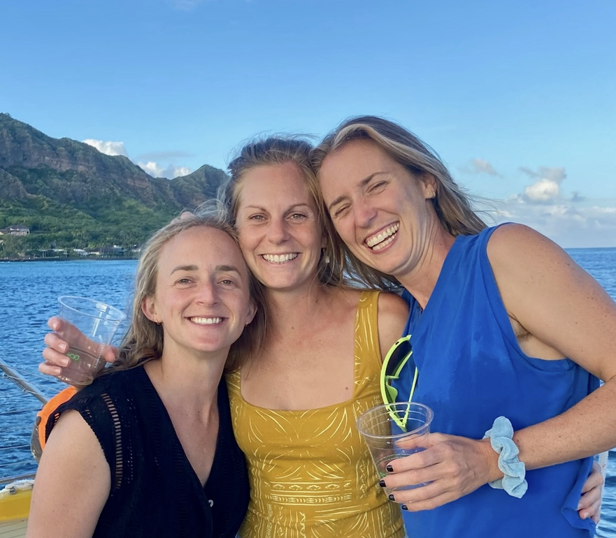 Three smiling women enjoying a boat trip with a mountain and ocean background.