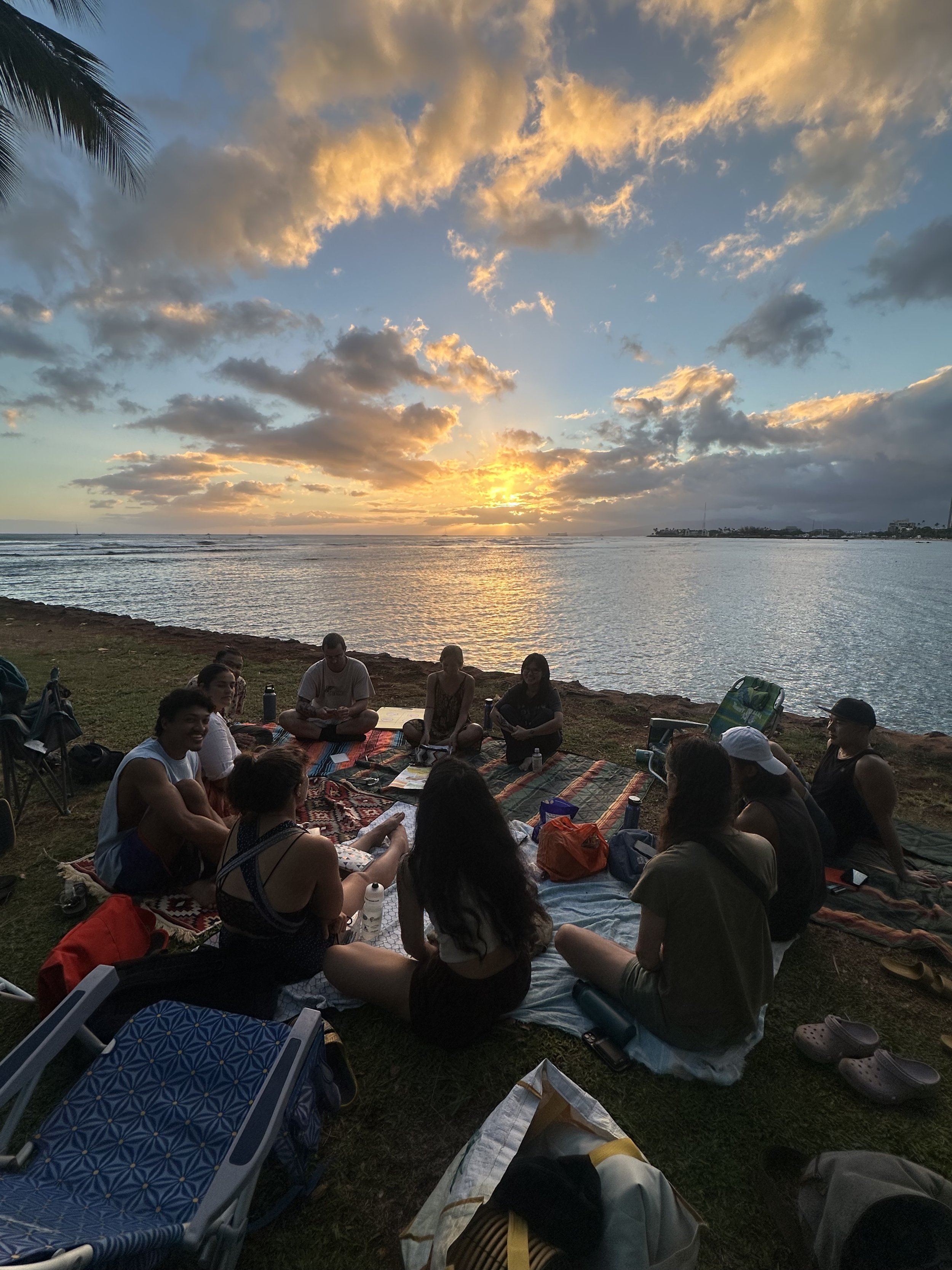 Group of people sitting on blankets and chairs by a body of water during sunset, with clouds in the sky and some boats in the distance.