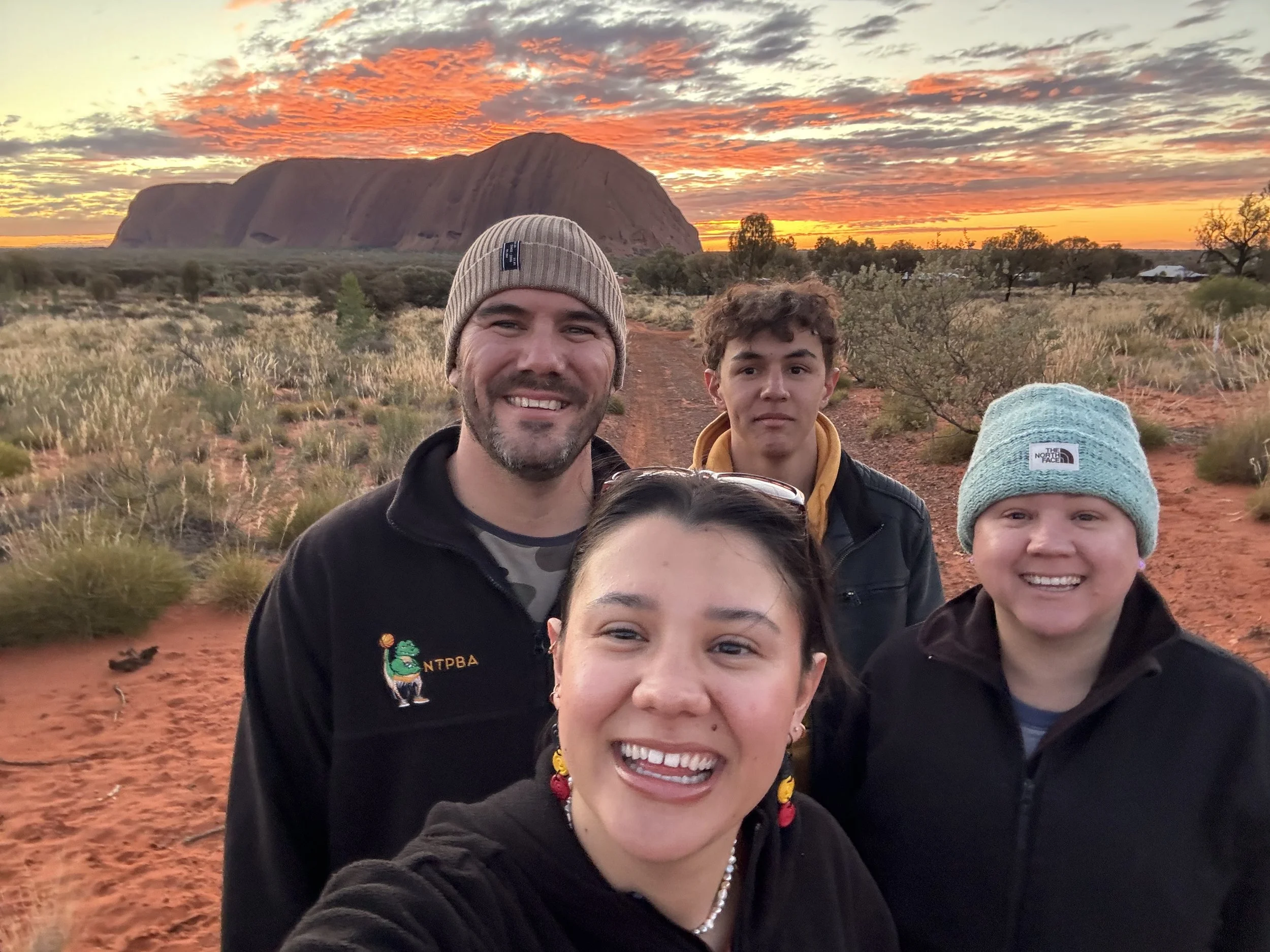 A group of four people smiling at the camera, with a scenic view of Uluru (Ayers Rock) at sunset in the background. The sky is filled with vibrant orange and yellow clouds. The landscape features red soil and sparse vegetation.