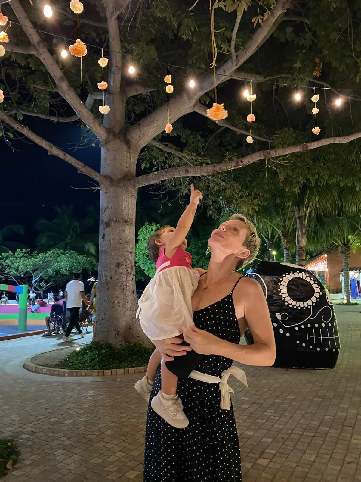 A woman holding a young girl outside at night, under a tree decorated with hanging string lights and paper lanterns, with a large painted black skull sculpture nearby, and people in the background.
